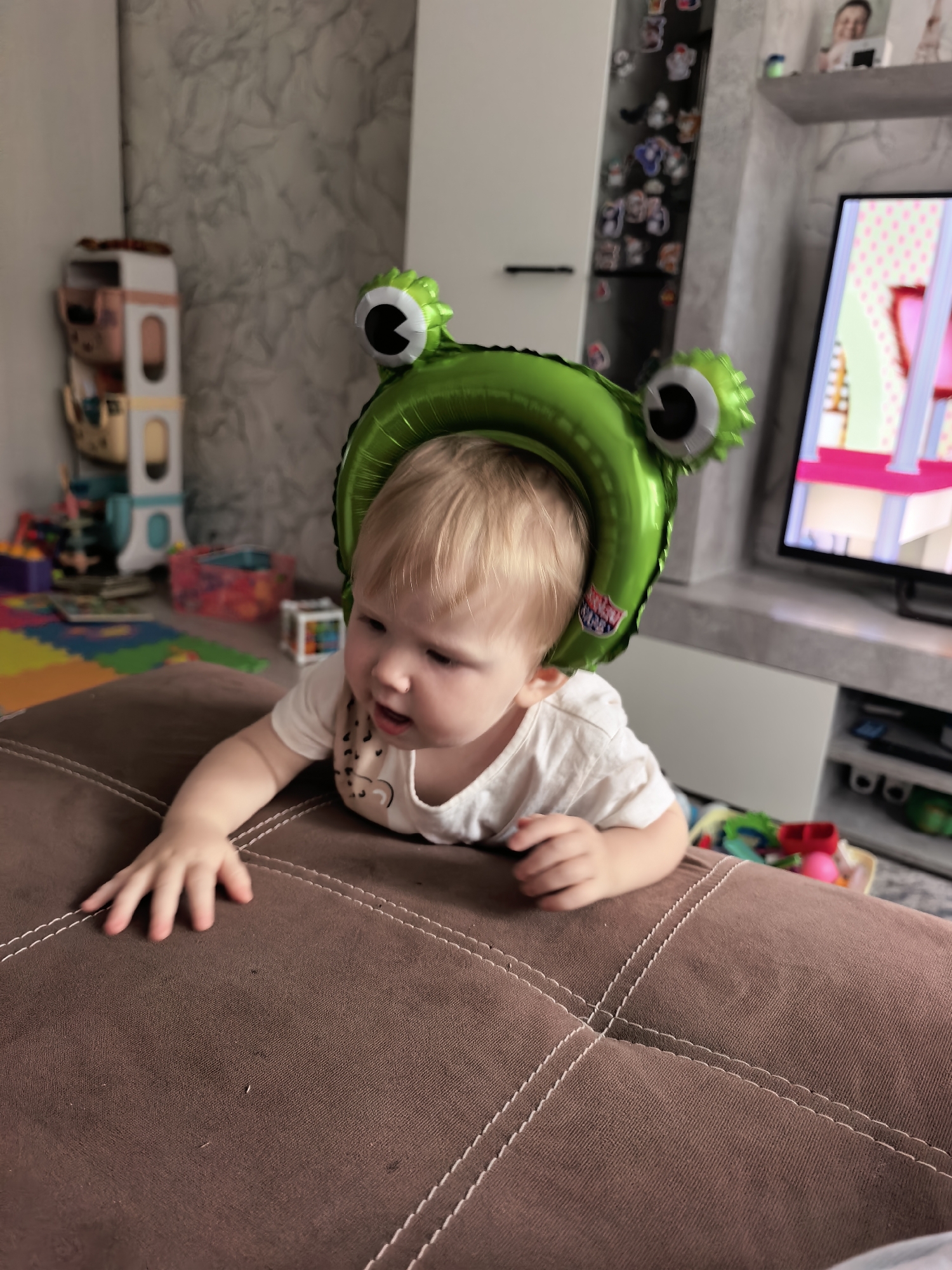 A blonde-haired baby wearing a frog-shaped balloon hat is lying on a brown couch. The baby is wearing a white t-shirt with a graphic design. The background shows a room with toys scattered on the floor, a shelf unit with various items, and a television displaying a cartoon. The scene appears to be indoors in a residential setting. The specific location within Sterlitamak, Russia, cannot be determined from the image.