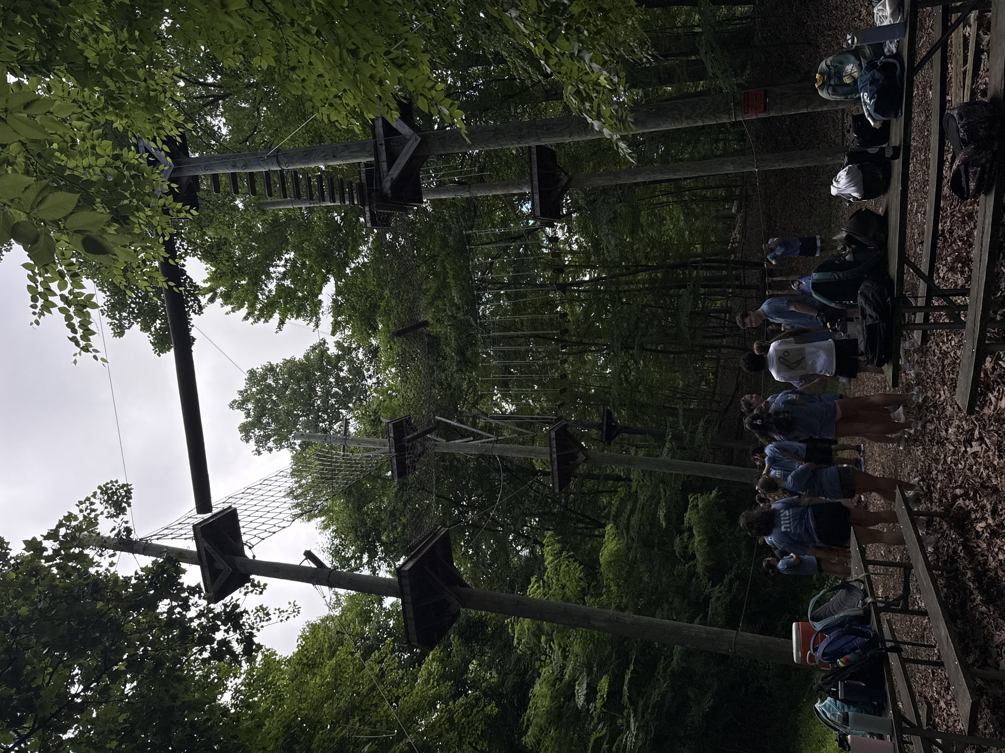 A group of teenagers stands around picnic tables laden with backpacks and coolers, in a wooded area with a high ropes course visible in the background. The ropes course consists of wooden platforms supported by tall poles, with various obstacles like nets, ladders, and suspended ropes connecting them. The teenagers, mostly wearing blue t-shirts, are gathered in small groups, some conversing while others appear to be waiting or observing. The ground is covered in fallen leaves, and the surrounding trees are lush and green, suggesting a summer or early autumn setting. The sky is overcast, casting a diffused light over the scene. The location is identified as Washington Township, United States of America, an area known for its outdoor recreational facilities.