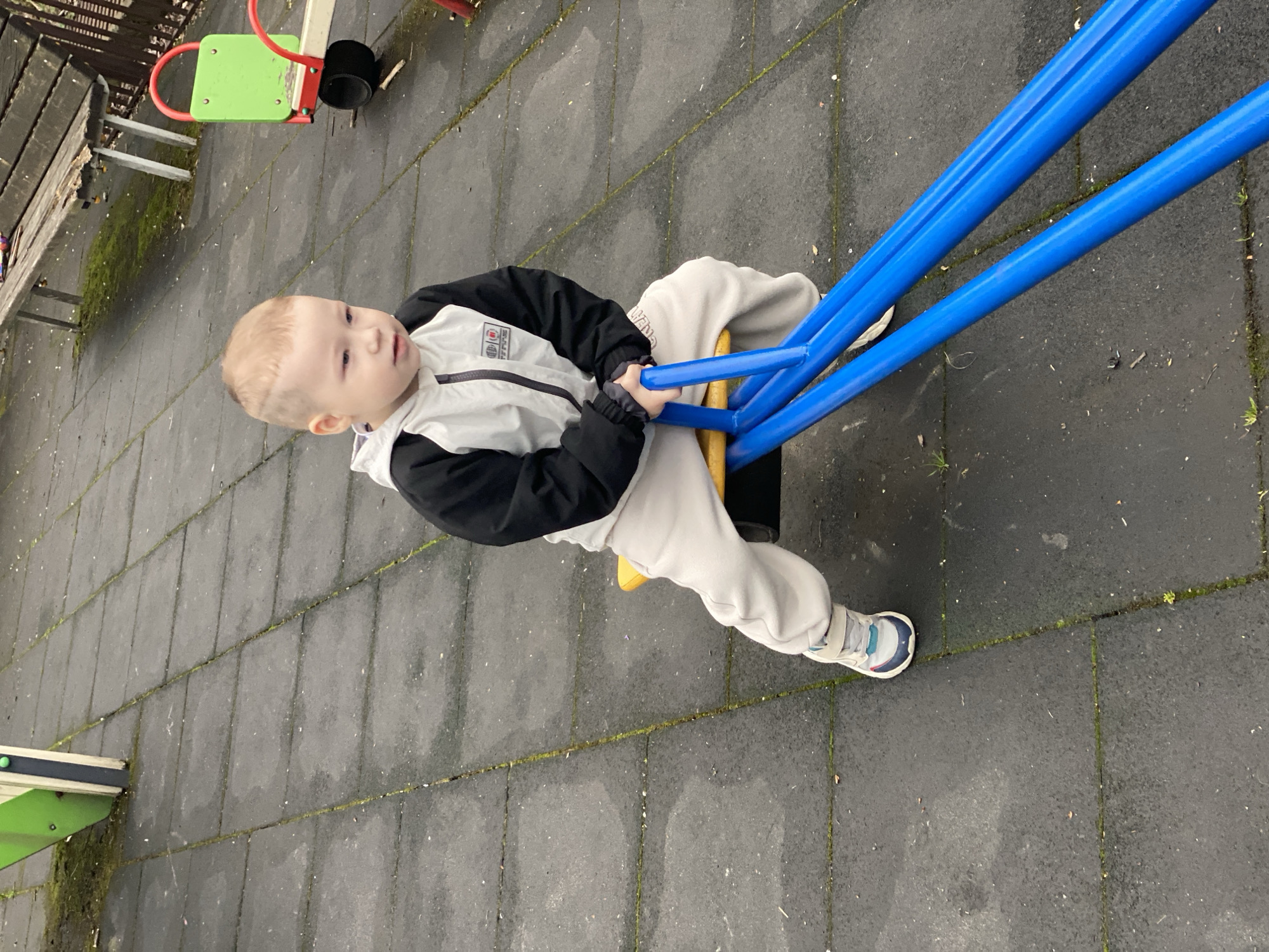 A young child is sitting on a blue seesaw in an outdoor playground. The playground has a dark gray surface. The child is wearing a light gray and black jacket, light gray pants, and sneakers. A green and red seesaw and a bench are visible in the background. The location is not identifiable as Sochi, Russia from the image alone.