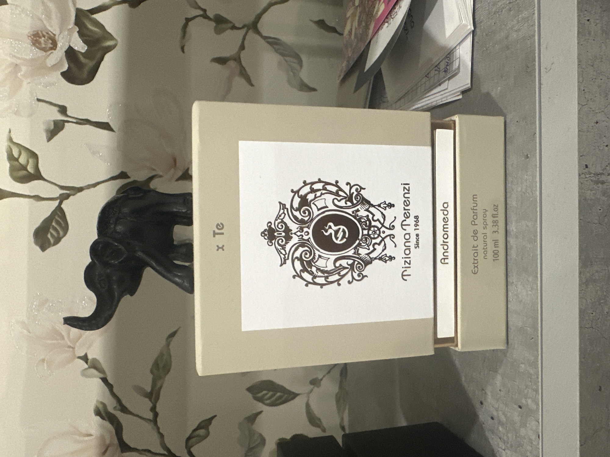 The image shows a perfume bottle box, labeled "Tiziana Terenzi Andromeda," with "Extrait de Parfum, natural spray, 100 ml 3.38 fl.oz" printed on it. The box is open, revealing the perfume bottle inside. The background features a floral wallpaper with a pattern of light pink and white flowers and green leaves. A dark elephant figurine is visible on the left side of the frame, partially obscuring the wallpaper. The scene appears to be indoors, likely on a shelf or table, with a grey, textured surface in the foreground and to the right. There are no people or animals other than the figurine visible. The lighting suggests it is daytime. The overall impression is one of a display of personal care items in a home setting.