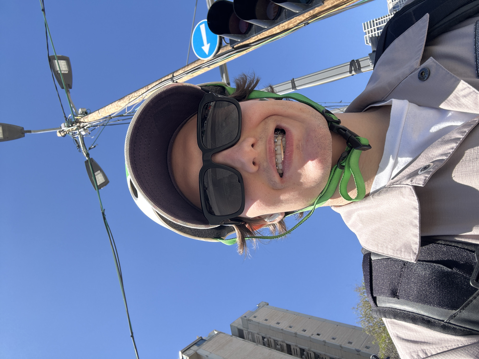 A low-angle, upward-facing selfie captures a smiling person outdoors on a bright, clear day in Givatayim, Israel. The person, visible from the chest up, wears black sunglasses, a light-colored helmet with vibrant green straps, and a light beige jacket over a white t-shirt. They have an open-mouthed smile, revealing braces on their upper teeth, and some stubble on their chin.
The background is a vivid blue sky, indicating sunny weather. Overhead, a utility pole with numerous cables is prominent, along with multiple streetlights. To the upper left of the person's head, dark traffic lights and a circular blue sign featuring a white downward-pointing arrow are visible. In the lower corners of the frame, parts of modern, light-colored buildings are seen. The image conveys a cheerful moment in an urban setting.