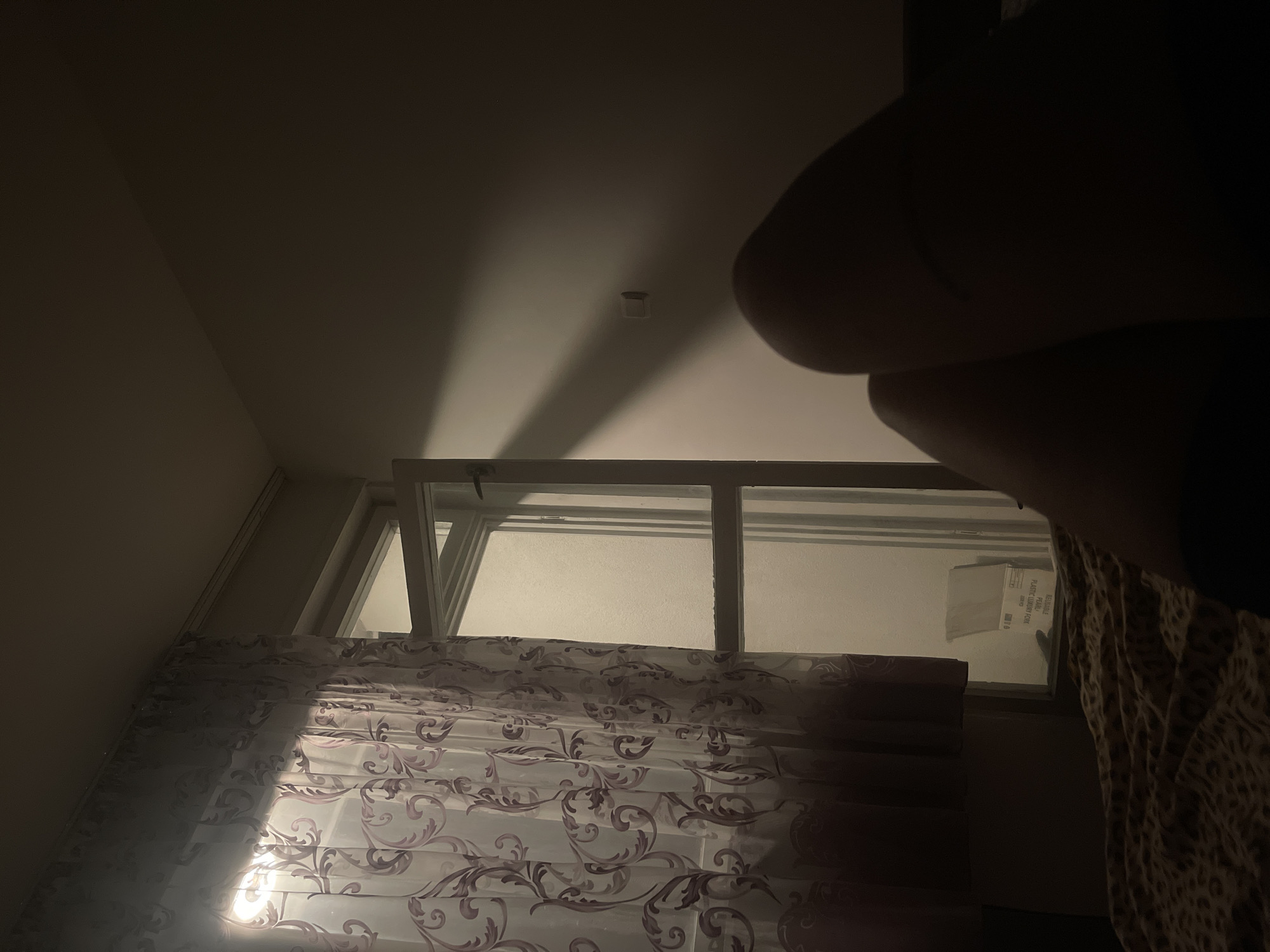 An indoor scene, possibly a bedroom, is dimly lit by light filtering through a sheer curtain with a purple floral pattern. The curtain covers a window or balcony door that is partially open. Outside the window, a light source illuminates a frosted glass panel and a white cardboard box. A strong shadow casts diagonally across the wall from the open door. In the foreground, a person's legs are visible, resting on a bed covered with a leopard-print blanket. The lower portion of the image is dark, obscuring the rest of the person and the immediate surroundings. The scene appears to be located in Varna, Bulgaria, suggesting a residential setting.