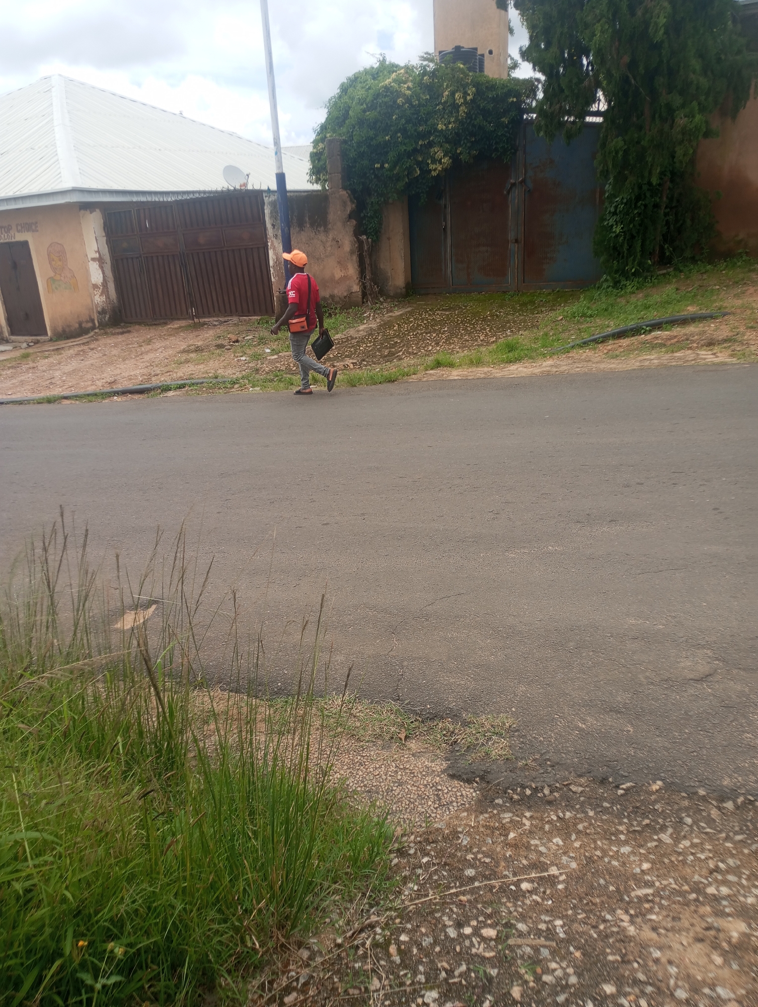 In Jos, Nigeria, a person wearing a red shirt, grey pants, and an orange cap walks along an asphalt road. The individual carries a dark bag over their shoulder and a smaller orange pouch across their chest. To the left of the road, a building with a white corrugated roof and a large brown gate is visible. To the right, a dense green tree and ivy-covered gates are present. The environment is a mixed urban and vegetated setting with a cloudy sky overhead.