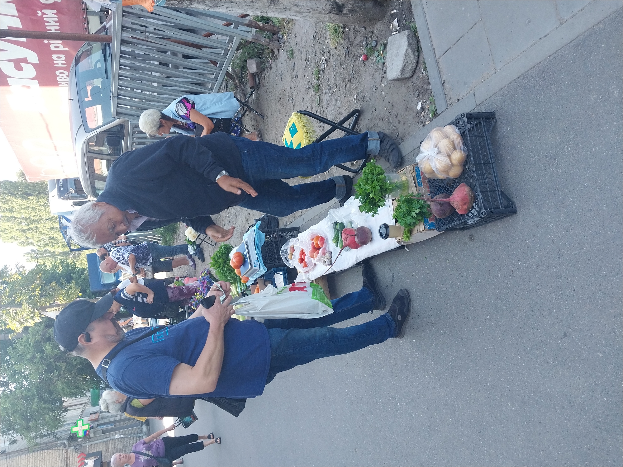 This is an outdoor street scene in Dnipro, Ukraine, likely a market setting. Two men are the primary subjects in the foreground, engaged in what appears to be a transaction. The man on the left, wearing a blue t-shirt and jeans, is holding a white plastic bag and looking down at something in his hands. The man on the right, wearing a dark jacket and jeans, is gesturing with his hands. They are standing next to a makeshift stall displaying various fresh produce, including tomatoes, cucumbers, beets, and potatoes. In the background, other people are visible, as well as parked vehicles and a building with a prominent sign. The environment suggests a bustling urban area with a focus on commerce.