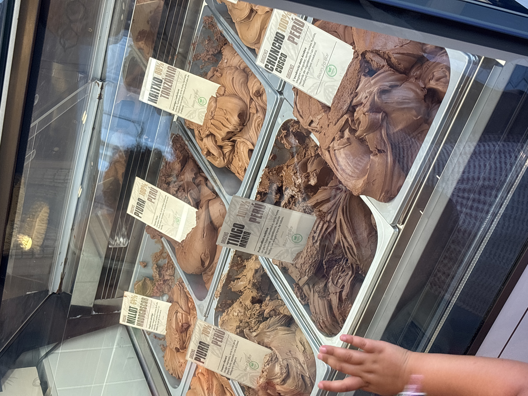 The image shows a close-up of a glass-fronted display case filled with various flavors of chocolate gelato or ice cream. Several small cards with text in French and Spanish are inserted into the gelato, indicating the origin and flavor. Some of the visible labels read: "MILLOT 64% MADAGASCAR", "PIURA 100% PERÚ", "KAITXO 80% TANZANIA", "TINGO 100% MARIA PERÚ", and "CHUNCHO 100% CUSCO PERÚ". The gelato appears to be a rich brown color with varying textures. In the foreground, the left hand of a young child is reaching towards the display case, suggesting a point-of-sale interaction. The overall environment appears to be an indoor retail space, likely a gelateria or artisanal chocolate shop. Based on the presence of Spanish text on the labels and the architectural style visible in the reflection on the glass (suggesting a European setting), this could be a shop in Palafrugell, Spain, a region known for its artisanal food products.