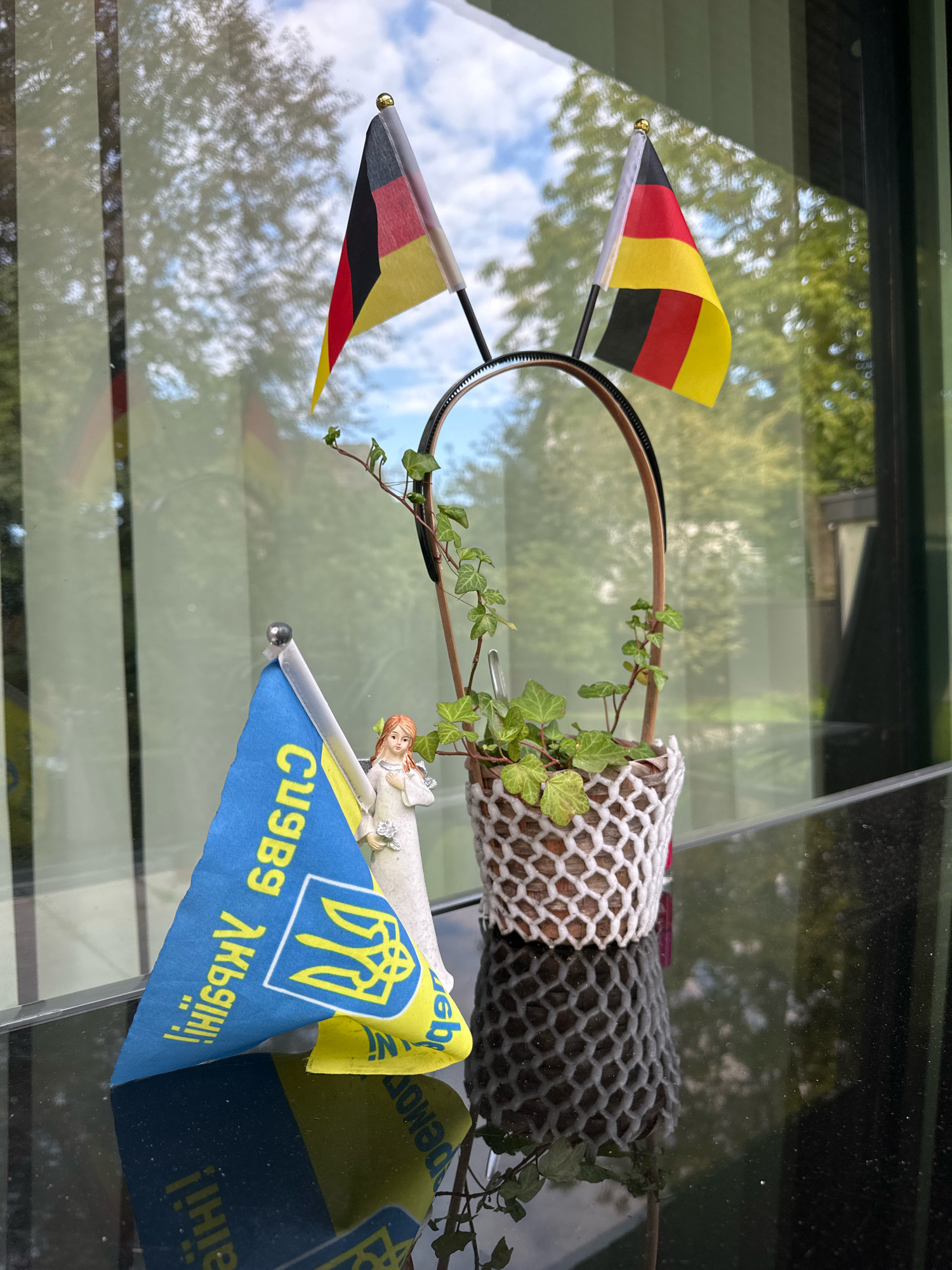 The image depicts a still life arrangement on a reflective black surface, likely a windowsill or table, set against a backdrop of a window with reflections of trees and a blue sky with clouds. Prominently featured are two small German flags on poles, attached to a black headband. To the left, a small figurine of a woman in a white dress holding flowers stands near a triangular blue and yellow flag, which is the flag of Ukraine. The Ukrainian flag has a crest and text in yellow on the blue background. A small potted plant with green ivy vines is positioned behind the flags and figurine, held within a woven, net-like decorative cover. The reflection on the black surface shows a distorted version of the objects and the surrounding environment. The scene suggests a display of solidarity or cultural connection. The location is identified as Krefeld, Germany.