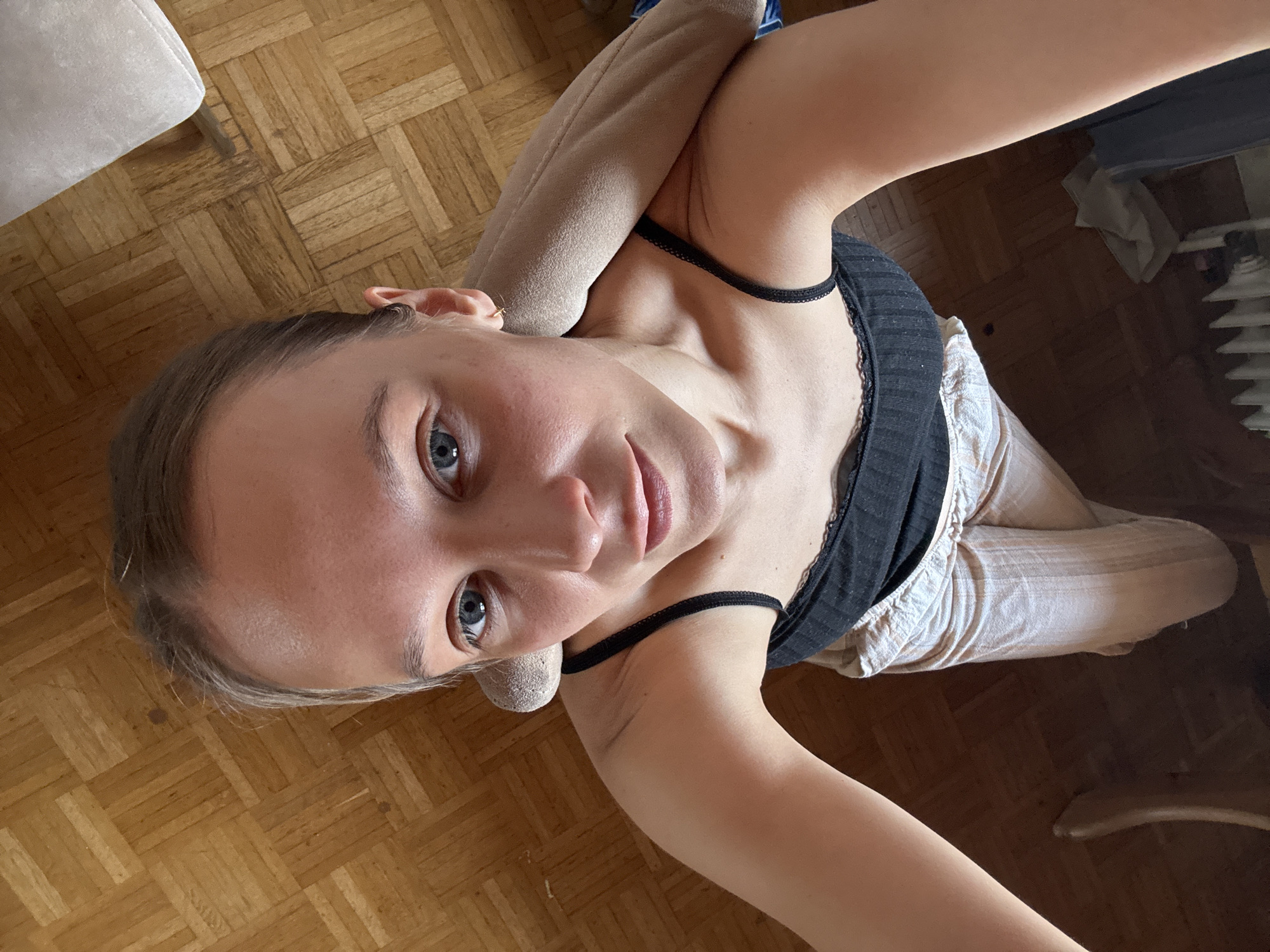 A young woman is pictured lying on a wooden floor. She is wearing a black tank top and light-colored striped pants. Her right arm is bent and resting on her head, supporting a beige stuffed animal. The room appears to be a living space, with a radiator visible in the background. The lighting suggests it could be daytime, possibly late afternoon due to the shadows. The woman's gaze is directed towards the camera, and she has a neutral expression. The floor is made of parquet wood, laid in a herringbone pattern. A white armchair is partially visible in the upper left corner of the frame. The setting appears to be indoors, in a home environment.