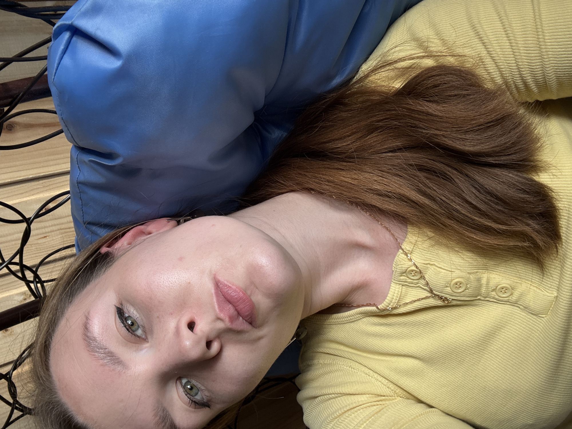 In a close-up, overhead shot, a young woman with long, reddish-brown hair and green eyes is captured. She is wearing a yellow, ribbed long-sleeved shirt with three buttons at the neckline and a delicate gold necklace. Her head is tilted, resting on a plump, royal blue cushion that appears to be part of outdoor furniture. The background features a wooden wall and some dark, curved metal elements, possibly part of a chair or hammock structure. The lighting suggests it is daytime. The image does not contain information that can be used to determine the specific location within the city of Italmas, Russia.