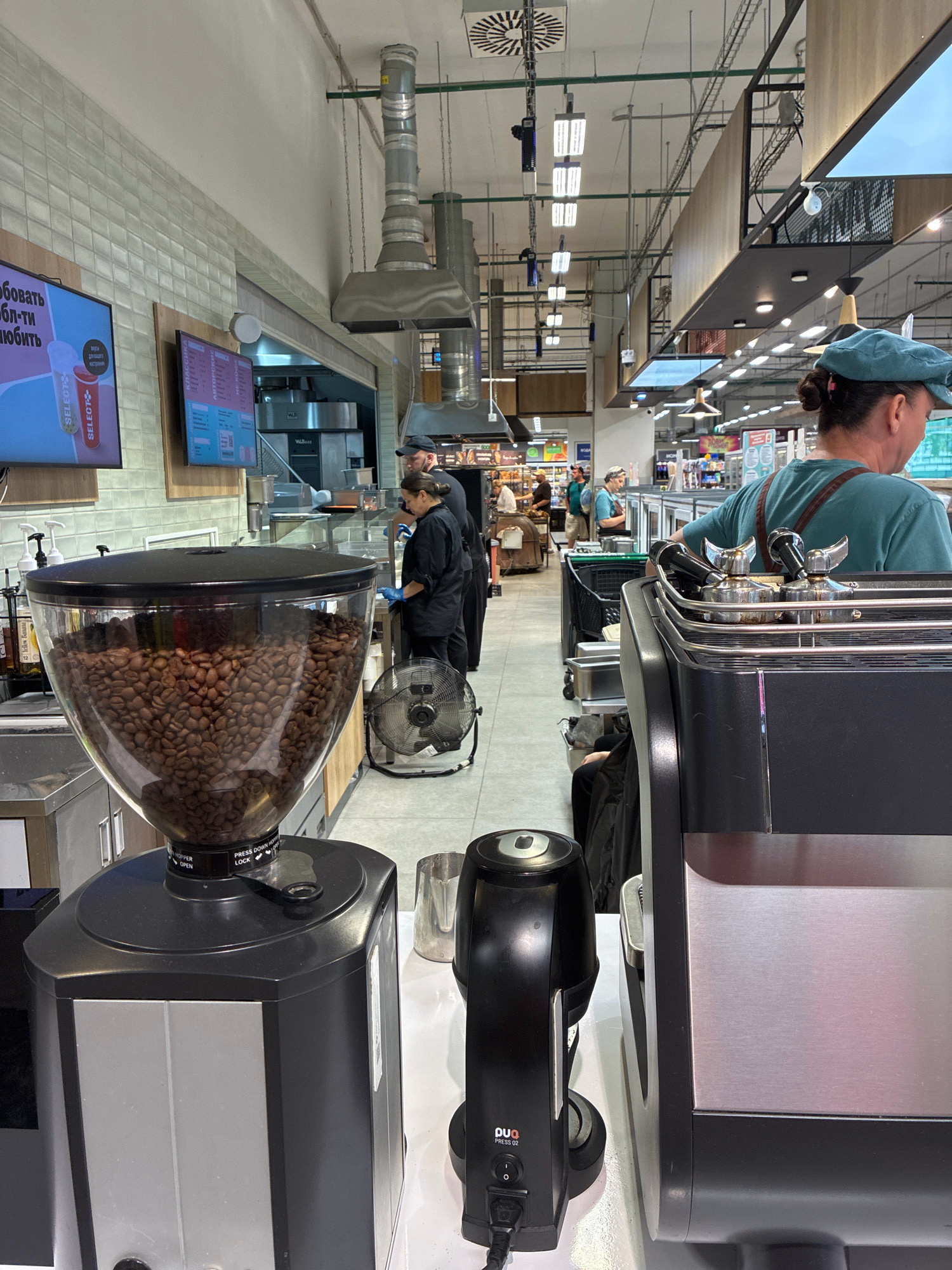 This image shows an interior view of a coffee stand within a supermarket, possibly in Sochi, Russia, given the Cyrillic text on a screen. In the foreground, a large coffee bean grinder is prominently displayed, filled with whole coffee beans. To its right is a stainless steel espresso machine with portafilters. A black tamper is visible in the center. A woman wearing a teal cap and apron is behind the espresso machine, facing away from the camera, and appears to be working. Behind her, a man in a black uniform is also present, along with another woman in a black uniform who is wearing blue gloves and interacting with a server station. A portable fan is situated behind the coffee grinder. In the background, shelves of products and other supermarket aisles are visible, along with overhead lighting and ventilation systems. A digital menu board with Cyrillic text is mounted on the wall to the left.