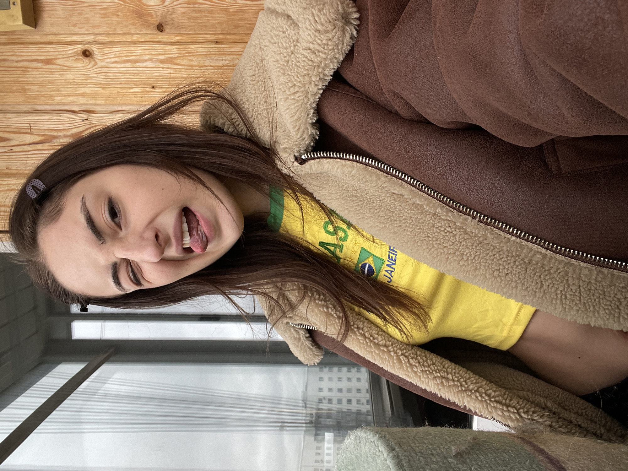 A young woman with long brown hair poses playfully for a photo indoors, sticking out her tongue and winking or squinting her left eye. She is positioned close to a window, which is largely obscured by horizontal blinds, revealing reflections of distant city buildings. Behind her is a light-colored wooden wall or paneling.
The woman is wearing a warm, brown faux shearling-lined jacket with a prominent zipper, open to reveal a bright yellow t-shirt underneath. The t-shirt features a graphic with the Brazilian flag and the word "BRAZIL" prominently, with "JANEIF" or "JANEIRO" partially visible below it. A small, dark-patterned hair clip is visible in her hair on the left side of her head. Her expression is jovial and mischievous.
The lighting appears to be natural daylight filtering in from the window. The heavy jacket suggests cool or cold weather outside. The setting is an urban indoor environment, specifically in Moscow, Russia, overlooking city infrastructure. In the foreground, a soft, light-colored fabric, possibly a blanket or rug, is visible.