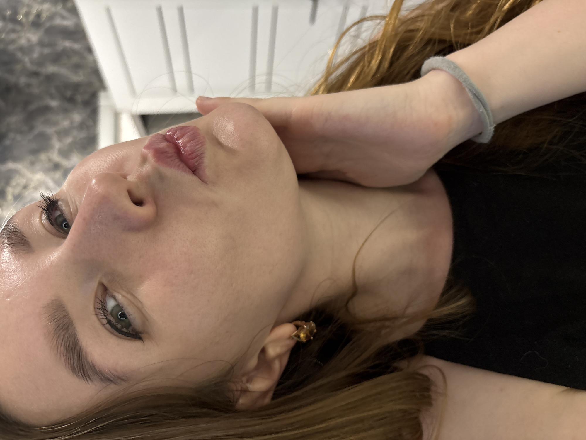 A close-up, slightly rotated, and partially obscured view of a woman with light skin and brown hair. She is wearing black clothing, and a grey bracelet is visible on her wrist. The woman has an earring in her left ear, and her facial features are prominent, including her eyes, eyebrows, nose, and pursed lips. A white, slatted object appears in the background. The location is not discernible from the image.