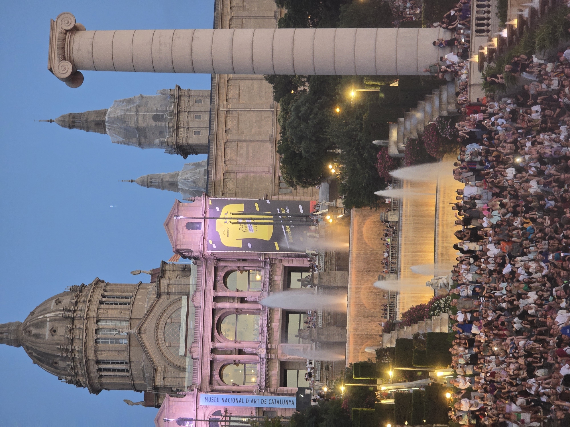 A large crowd of people are gathered on a tiered seating area, facing a grand building. The building, which appears to be the National Palace of Catalonia, is illuminated in pink and features ornate architecture with a large dome and several smaller towers. A massive column stands to the right of the building. In front of the building, a multi-tiered fountain is active, with water cascading down. The scene is set at dusk, with the sky a deep blue. Banners are hung on the building, one of which is yellow with text that is partially obscured by the fountain. The crowd is diverse in age and attire, with many individuals holding up phones, suggesting they are capturing the moment. The overall atmosphere appears to be one of a public event or performance.