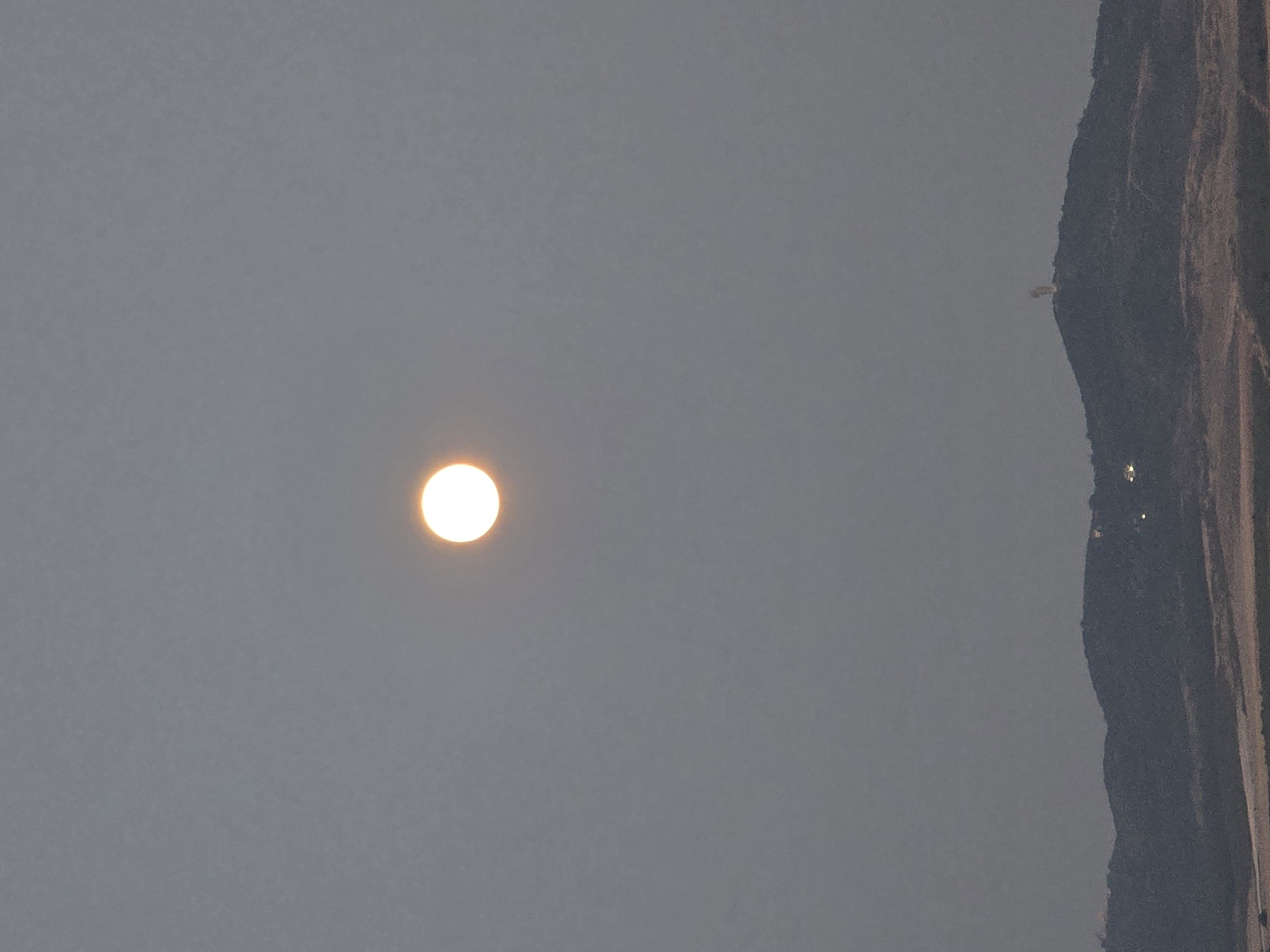 The image captures a twilight scene with a full moon prominently displayed in the center of the dusky sky. Below the moon, a silhouetted mountain range dominates the lower half of the frame. On the peak of one of the mountains, a statue of Saint George is visible, a well-known landmark in the city of Yambol, Bulgaria. Scattered lights can be seen in the distance on the mountainside, suggesting the presence of a settlement. The overall atmosphere is serene and slightly hazy, typical of dusk.
