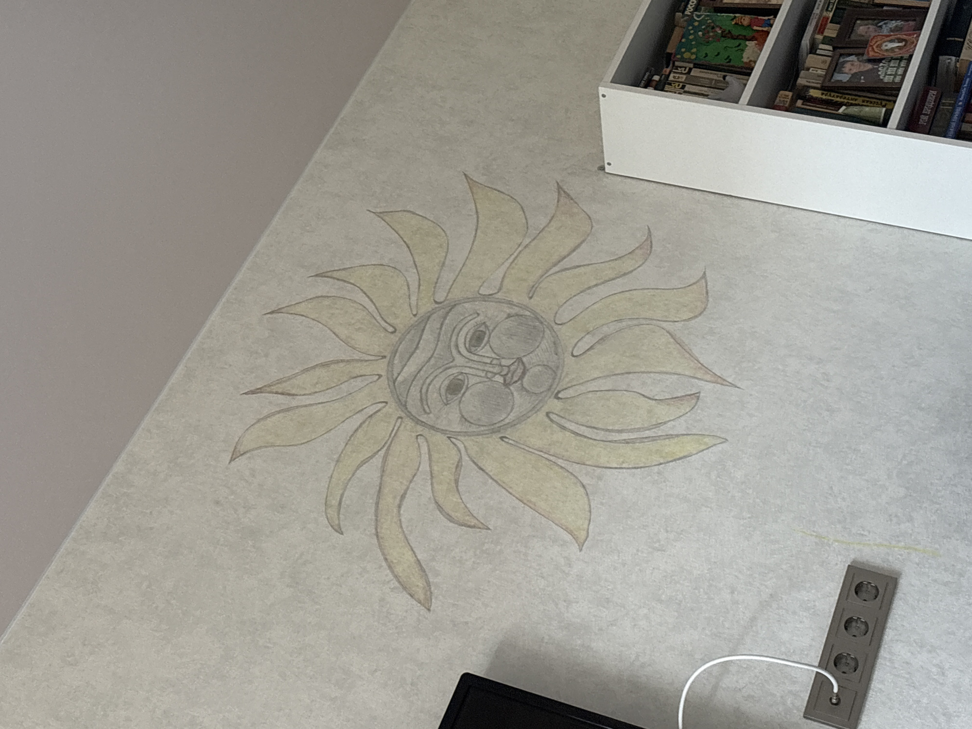 The image shows a wall with a drawing of a sun and a bookshelf. The sun drawing is in yellow and gray and is on the left side of the wall. The bookshelf is on the right side of the wall and is filled with books and pictures. A power strip is visible on the wall below the sun drawing. The overall scene suggests a domestic indoor environment.