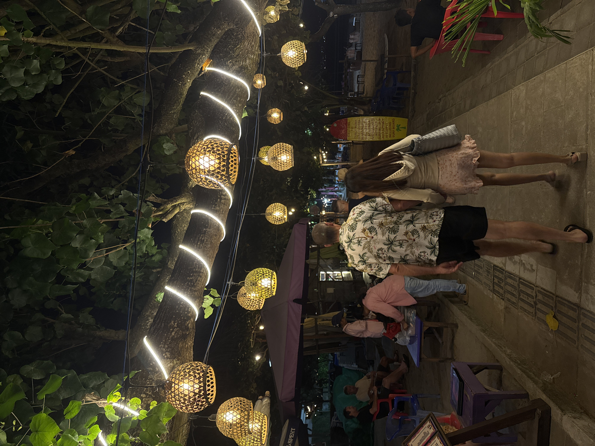 An outdoor nighttime scene is depicted in Kuta, Indonesia, featuring a pathway lined with restaurants and beachside establishments. A prominent feature is a large tree adorned with numerous woven, spherical pendant lights and a string of white LED lights that create a warm, ambient glow. In the foreground, a couple walks away from the camera. The male is wearing a floral short-sleeved shirt and black shorts, while the female is wearing a light-colored hooded sweatshirt and a pink floral skirt. Behind them, several other individuals are visible. Some are seated at tables, while others are standing near food stalls or umbrellas. Tables and chairs are set up in front of establishments, and a surfboard with surf school information is displayed on the right side of the pathway. The ground is paved with tiles, and a yellow leaf is visible on the pathway. The atmosphere suggests a lively and popular tourist area at night.
