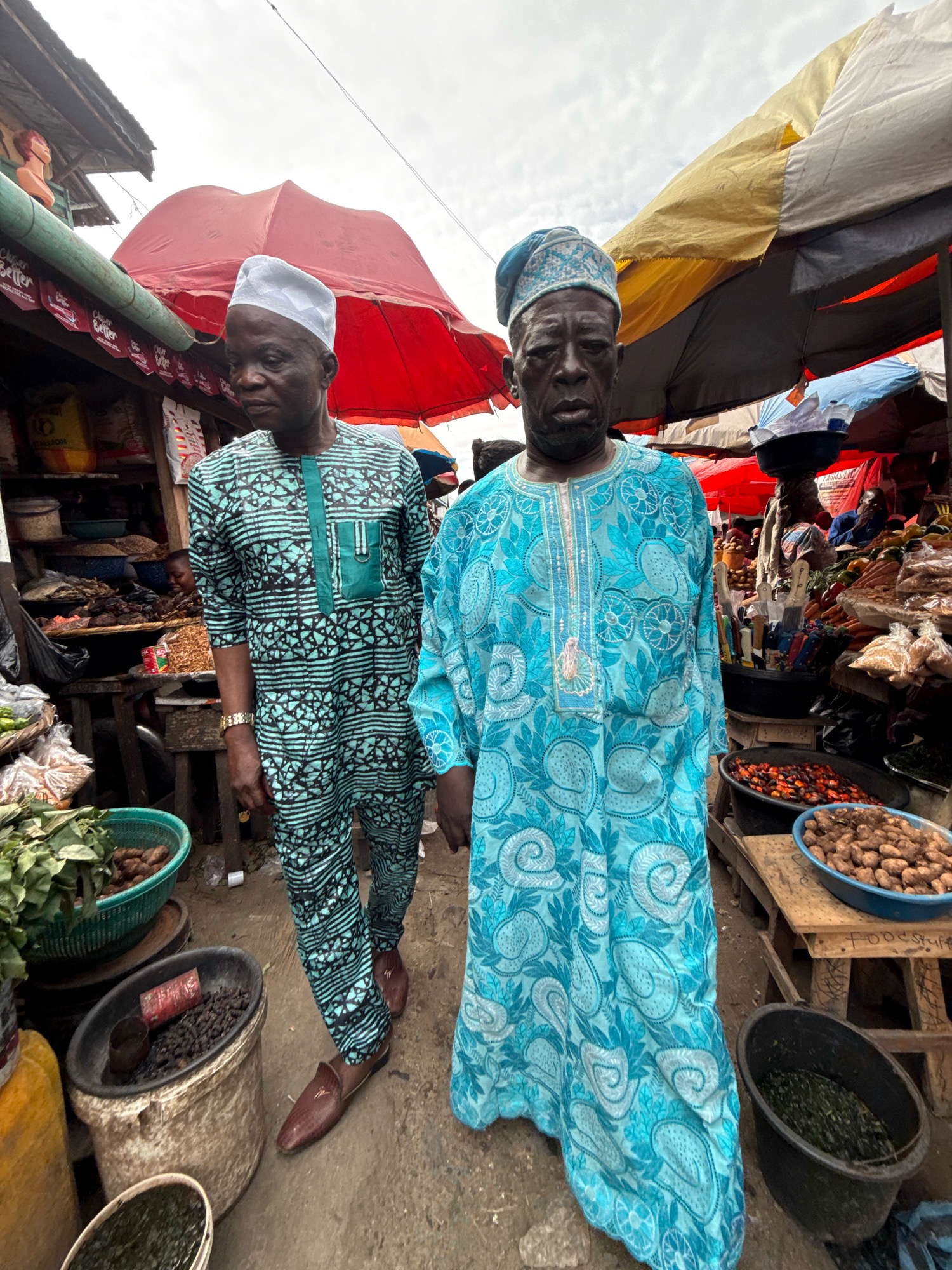 Two men, dressed in traditional African attire, stand in a bustling open-air market in Lagos, Nigeria. The man on the left wears a teal and black patterned tunic and trousers, with a white cap. The man on the right is clad in a light blue, intricately patterned kaftan and a matching blue patterned cap. They are positioned in the foreground, facing forward, with the market stalls and goods forming the backdrop.
The market environment is characterized by a variety of goods displayed on wooden tables and in baskets. These include what appear to be dried fish, vegetables, spices, and root vegetables. Several colorful awnings and umbrellas, predominantly red and yellow, are visible overhead, providing shade. The ground is dusty and uneven, typical of a busy marketplace.
The man on the left has a neutral expression, while the man on the right's expression is more serious, with his gaze directed towards the camera. Their attire and the surrounding market suggest a cultural and commercial setting in Lagos. The overall scene captures a moment in the daily life of this vibrant Nigerian city.