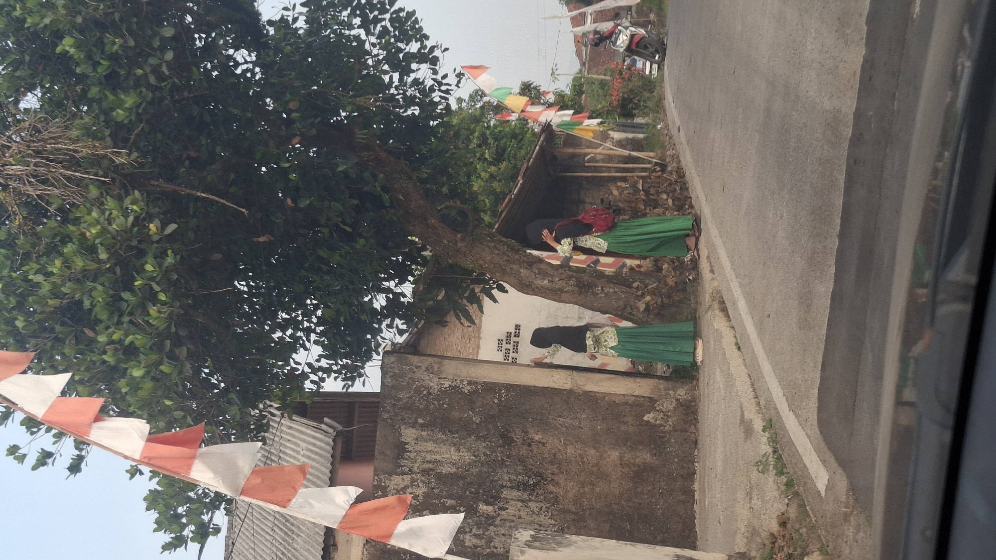 This image depicts a street scene in Cibinong Hilir, Indonesia, featuring two young females in green skirts and headscarves standing near a large tree. One girl is reaching towards a wall, while the other is holding her headscarf. Red and white flag bunting is visible along the street, attached to a building and a tree. A motorcycle is parked on the right side of the road. The setting appears to be a rural or semi-urban area with a rough concrete wall, a dirt path, and asphalt road. The foliage of a large tree dominates the upper portion of the image.