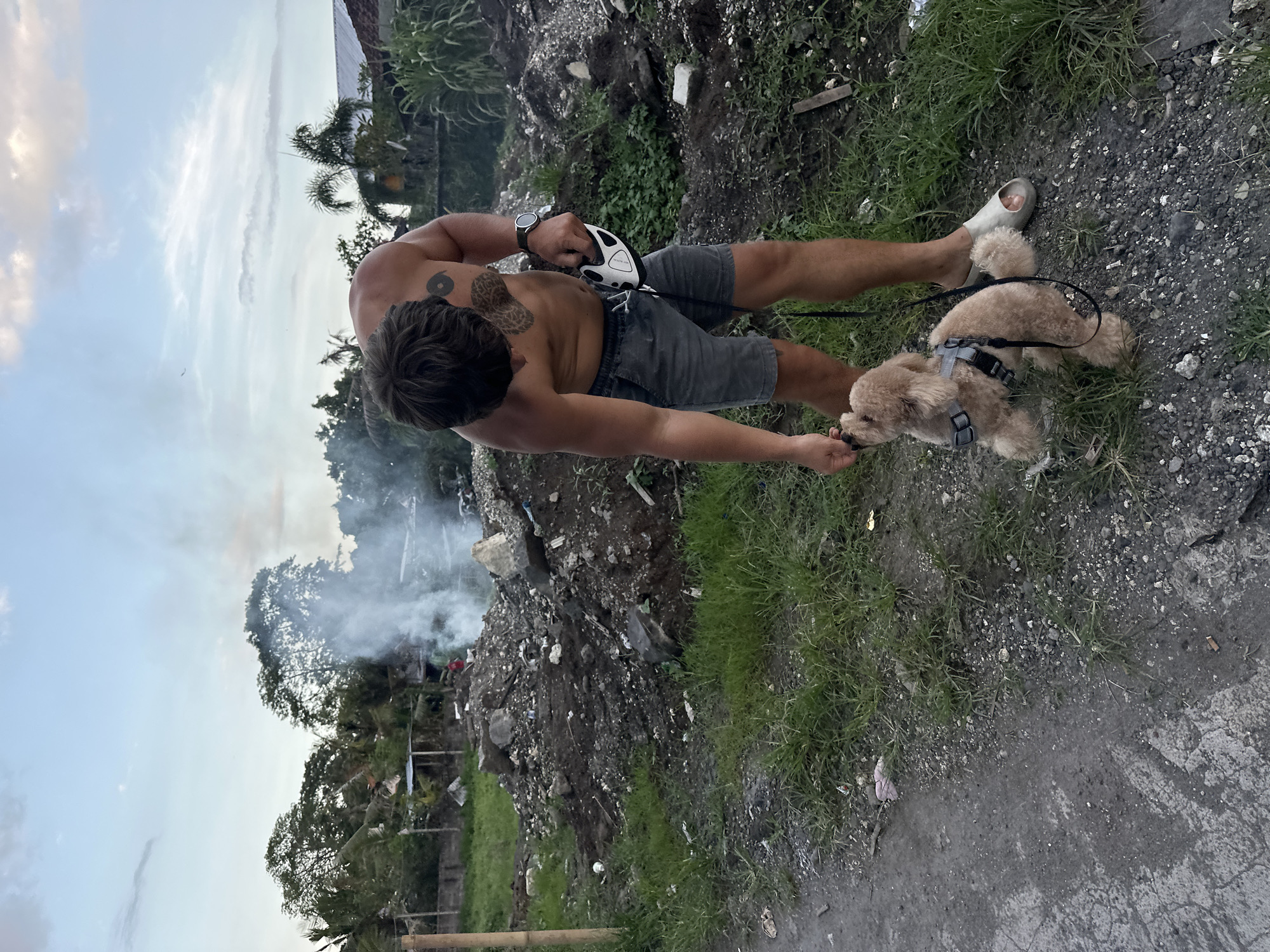 A shirtless man in North Kuta, Indonesia, wearing shorts and one sandal, is crouched down near a dog. The man has tattoos on his torso and arm, and a watch on his wrist. He appears to be interacting with the small, brown dog on a leash. In the background, there is a trash heap and smoke rising near trees. The sky is visible in the upper left of the frame. The ground is a mix of dirt, grass, and gravel.
