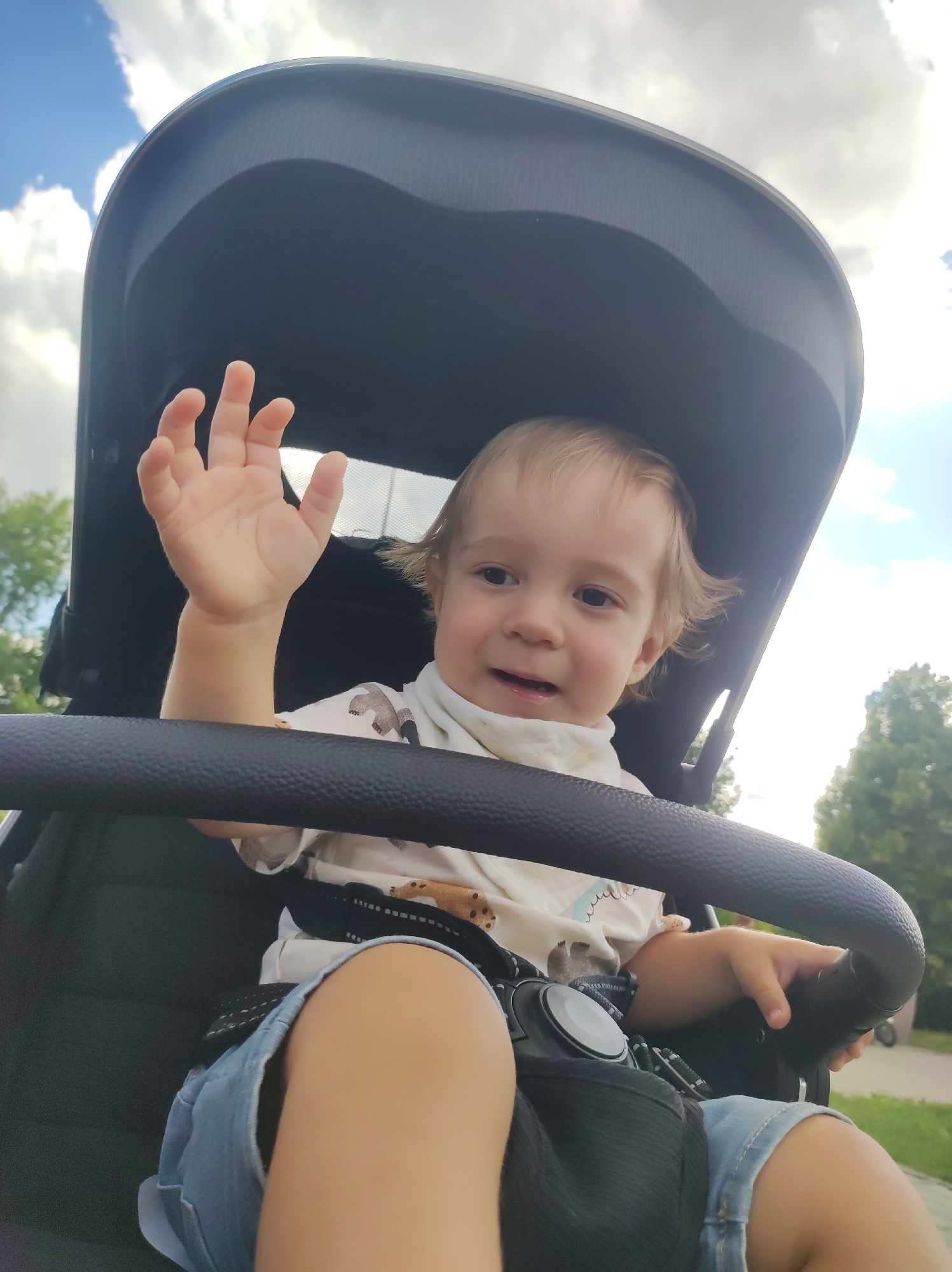 A young child, likely a toddler, is sitting in a stroller outdoors. The child has blonde hair and is looking towards the camera with a slight smile. Their right hand is raised, palm open, as if waving. The child is wearing a light-colored t-shirt with a pattern and denim shorts. The stroller has a dark canopy and a black handlebar in the foreground. The setting appears to be a park or outdoor recreational area, with green trees and a bright sky with clouds visible in the background. The lighting suggests it is daytime, possibly late morning or early afternoon. There is no visible text in the image. The overall mood is cheerful and candid, capturing a moment of a child's outing.