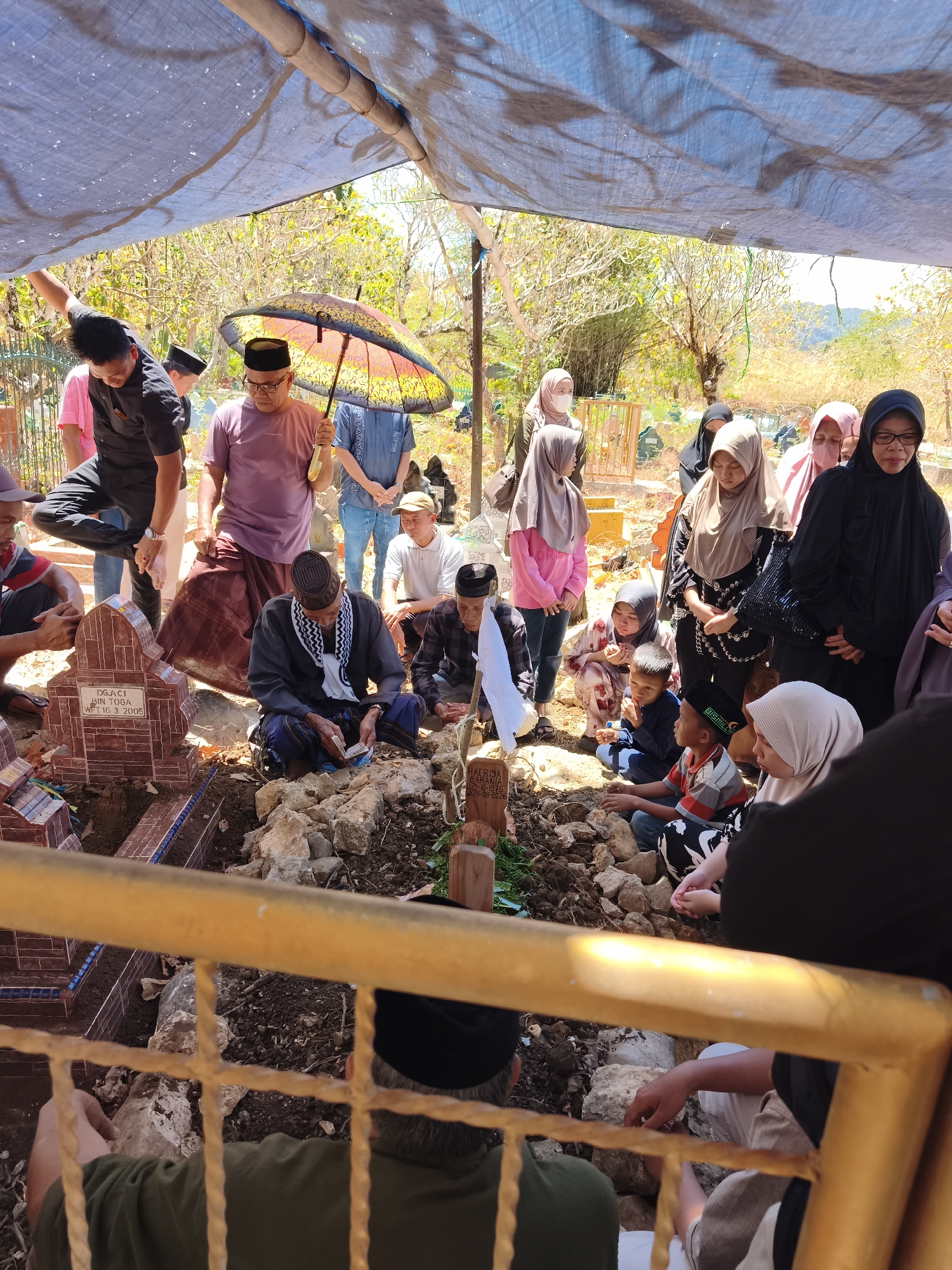 An outdoor, overhead view captures a gathering of people at a grave in Tonasa, Indonesia. A blue tarp is draped over a bamboo structure, providing shade for the mourners. At the center of the scene, a man in a black skullcap and patterned sarong kneels by a grave, reading from a book. Several other individuals, dressed in various modest attire including hijabs and skullcaps, are positioned around the gravesite, some sitting and others standing. The graves themselves are marked with small headstones and are surrounded by rocks and dirt. The environment appears dry and sun-drenched, with sparse vegetation and trees visible in the background. A yellow metal railing frames the foreground, partially obscuring the lower portion of the image.