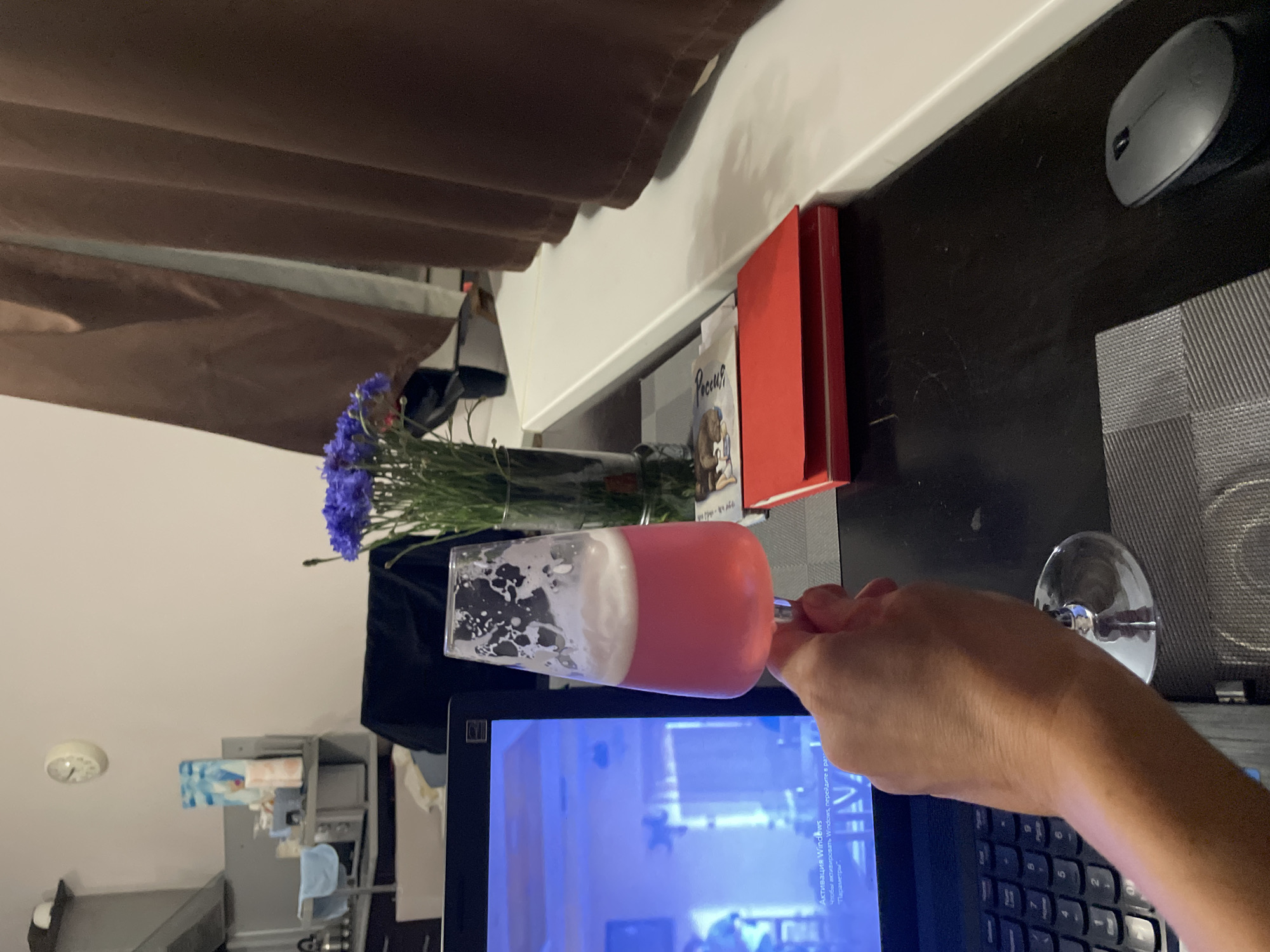The image depicts a close-up view of a person's hand holding a stemmed glass filled with a pink beverage and foam, positioned in front of a laptop screen displaying a blue interface with Russian text. The scene is set indoors, likely in a domestic environment. A vase of purple flowers is visible in the background, alongside a stack of red books and a computer mouse on a dark desk. The overall impression is one of relaxation and personal time, possibly in a home office or living area. The location is specified as the city of Sochi, Russia.