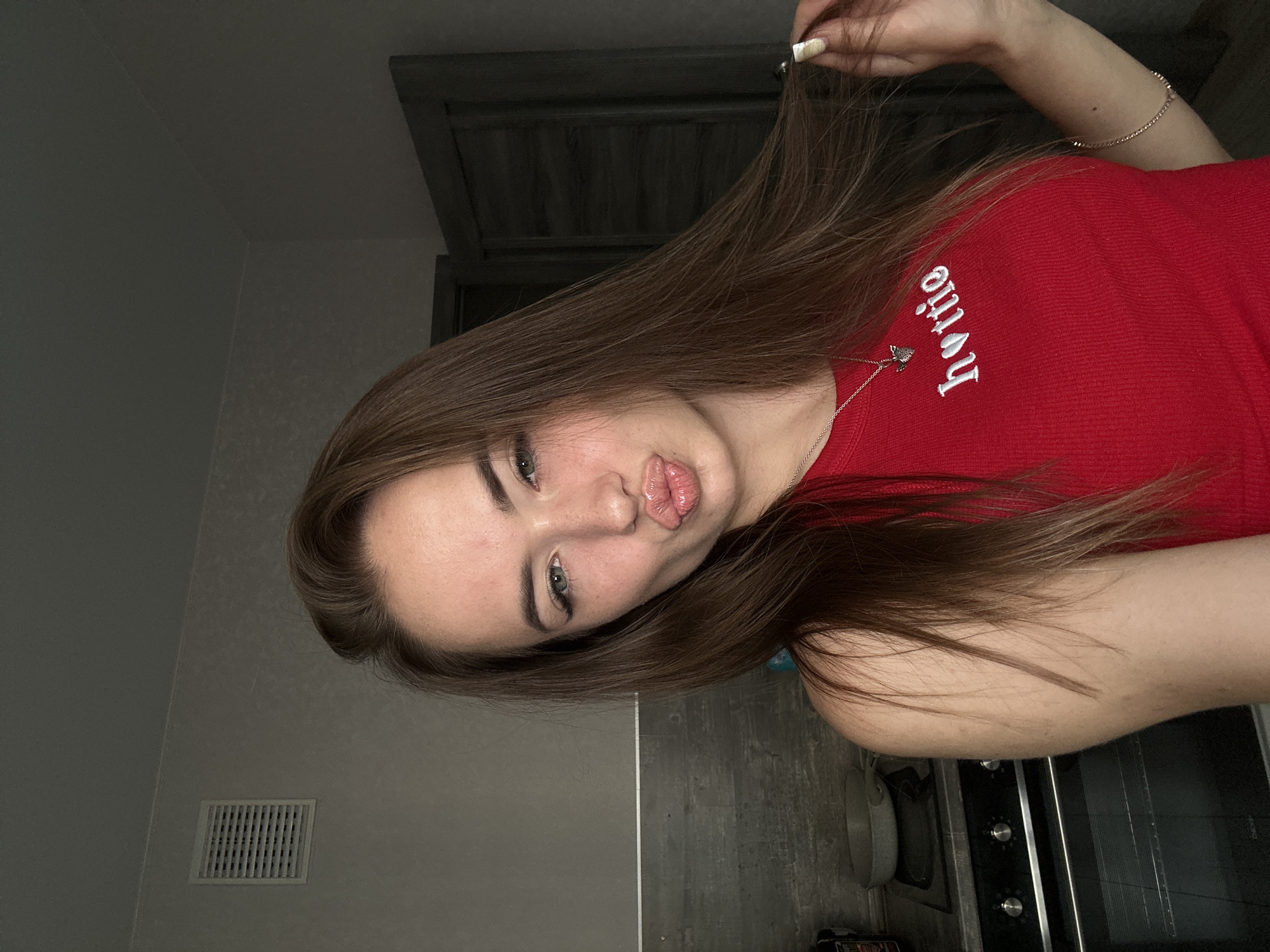 The image depicts a young woman in a close-up selfie, with her head and upper torso filling the frame. She has long, brown hair that cascades over her shoulders and down her chest. Her eyes are a light color, and she is wearing subtle eye makeup, including winged eyeliner. Her lips are pursed in a kissing pose, and she has a smattering of freckles across her nose and cheeks. She is wearing a red, ribbed tank top with the word "hottie" embroidered on it in white, with a small heart icon replacing the "o". A delicate silver necklace with a small charm is visible around her neck. On her left wrist, she wears a thin silver bracelet.
She is positioned in what appears to be a domestic setting, likely a kitchen, as evidenced by a black oven with control knobs visible in the background to her left. Above the oven, there is a grey tiled backsplash. To her right, a dark grey door is partially visible. The lighting suggests an indoor environment, possibly with natural light coming from the side, creating some shadows. The overall impression is casual and personal, typical of a social media selfie. The location is not specified beyond "Unknown, Unknown."