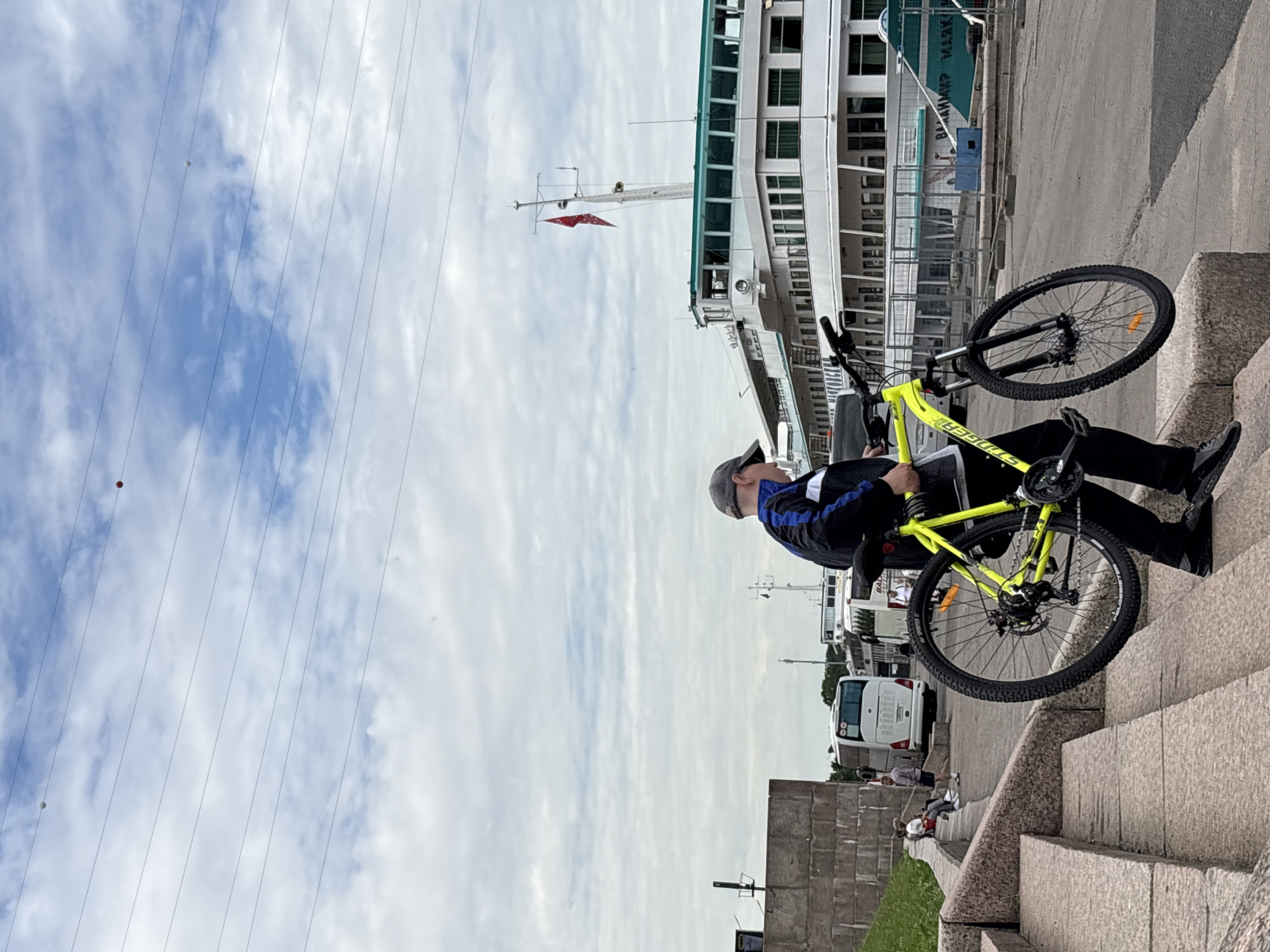 The image depicts a person carrying a bright yellow bicycle up a set of stone stairs. The individual is wearing a dark jacket with blue stripes and a grey baseball cap. In the background, a large white ship with green trim is docked, and several white buses are visible. The scene appears to be in an urban environment, possibly a port or waterfront area. The sky is cloudy with visible power lines strung across it, one of which has two small spherical objects hanging from it. The specific location of Shushary, Russia, is not directly indicated by any visible landmarks or text in the image.