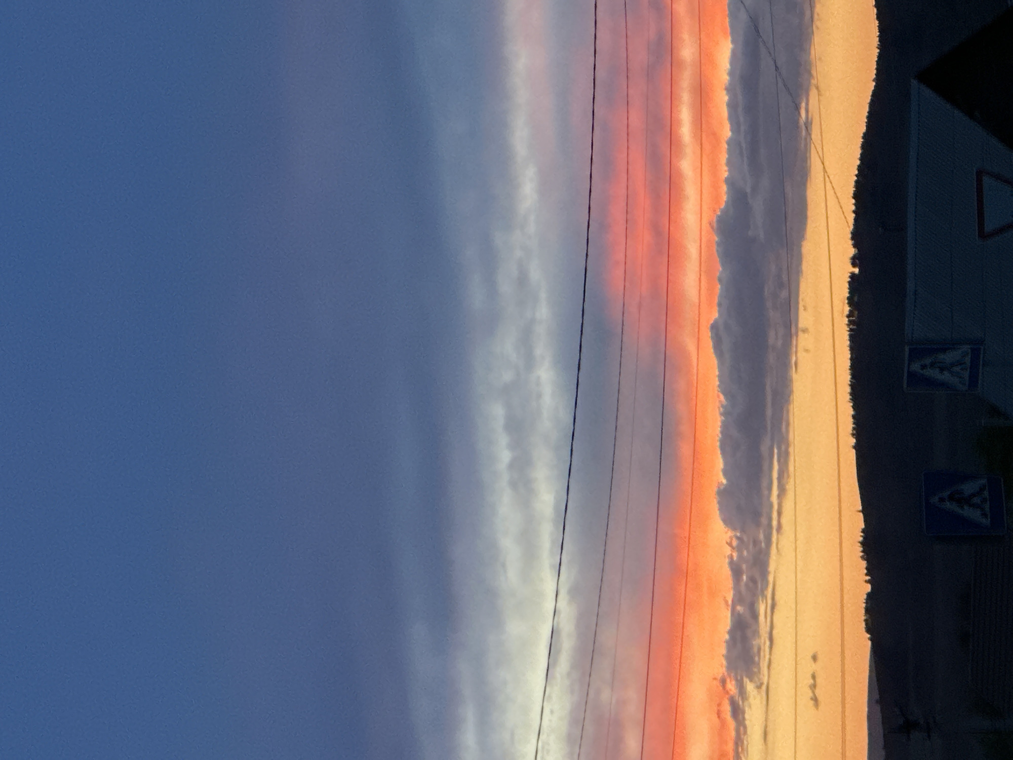 The image depicts a sunset over Uchaly, Russia. The sky transitions from a deep blue on the left to a vibrant orange and yellow on the right, with streaks of clouds running vertically. Power lines traverse the sky. A dark silhouette on the right suggests trees and buildings, including traffic signs.