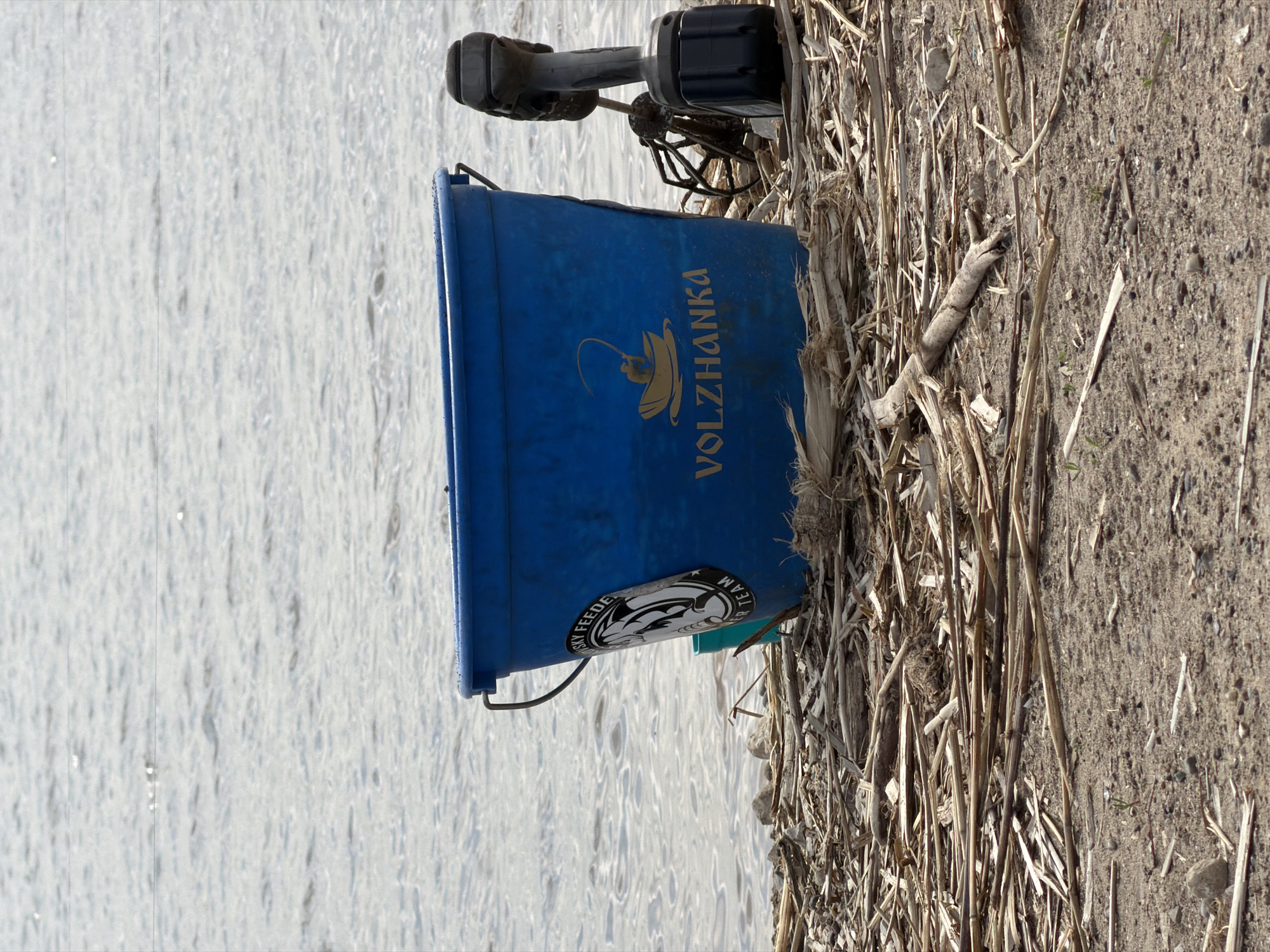 The image depicts a blue Volzhanka fishing bucket and fishing equipment on a riverbank near Krasnyi Bor, Russia. The bucket is positioned adjacent to the water. The ground is covered with dry reeds and earth.