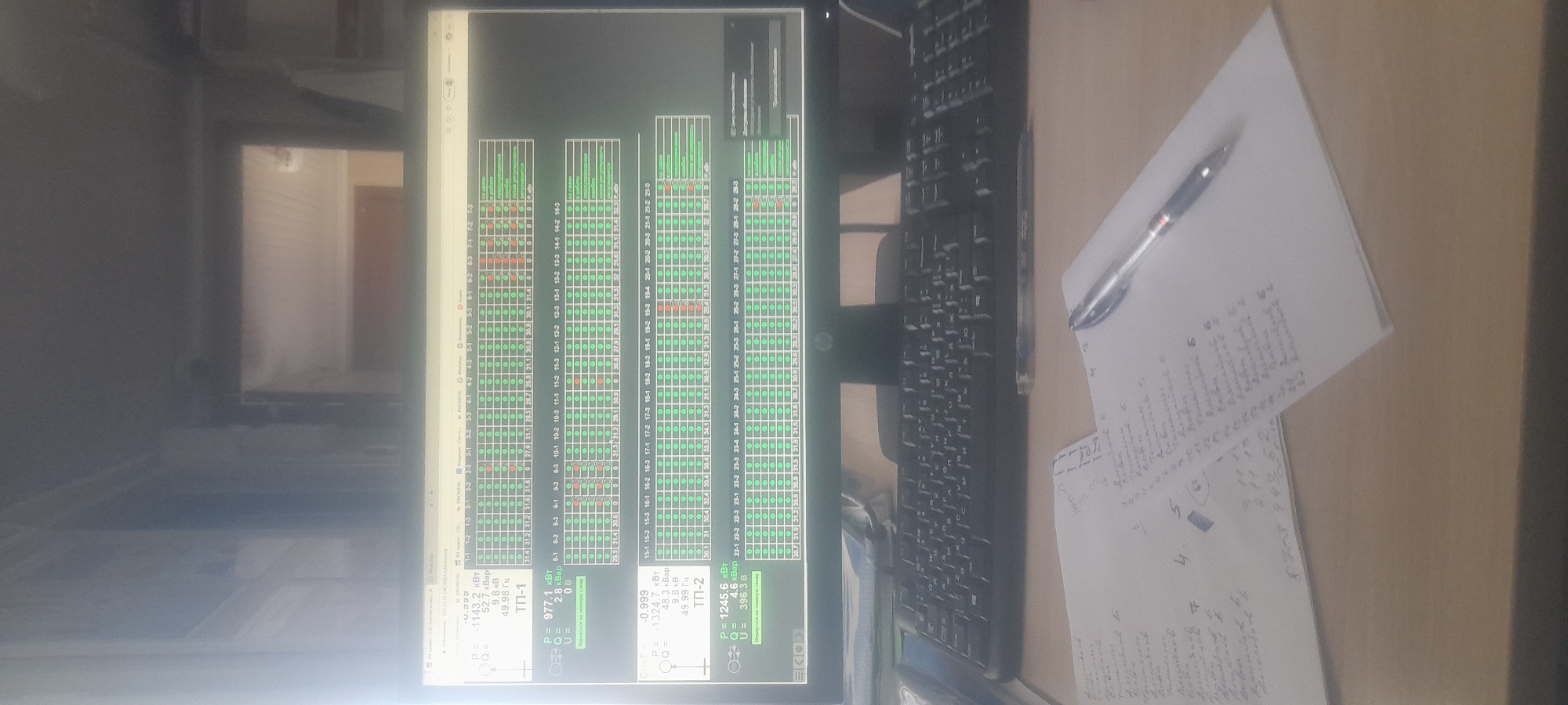 This media file depicts a close-up, slightly angled shot of a computer monitor displaying technical data, likely in an industrial or control room setting. The screen shows a grid of green and red circles, representing status indicators, alongside numerical data presented in rows and columns. There are labels indicating "ТП-1" and "ТП-2," which could refer to technical parameters or substations. Textual elements on the screen include values for "P" (power), "Q" (reactive power), "U" (voltage), and frequencies in Hertz (Гц) and Kilovolt-Ampere reactive (кВар). Some of the numerical values are large, such as 1245.6 kW and -1324.7 kW.
To the right of the monitor, a black keyboard is visible, suggesting a user interaction point. A pen rests on a piece of paper with handwritten notes, further indicating an active work environment. The paper contains a mix of numbers and illegible script, possibly related to the data displayed on the screen or a separate task.
The scene appears to be indoors, with the monitor and keyboard placed on a desk. The lighting is somewhat dim, with reflections visible on the monitor screen, hinting at the presence of other light sources in the room. The overall impression is of a functional workspace focused on monitoring and managing technical systems. The specific location context of Ushtobe, Kazakhstan, combined with the technical data on the screen, suggests this could be a control room at a power substation or industrial facility in that region. There are no people visible in the image.