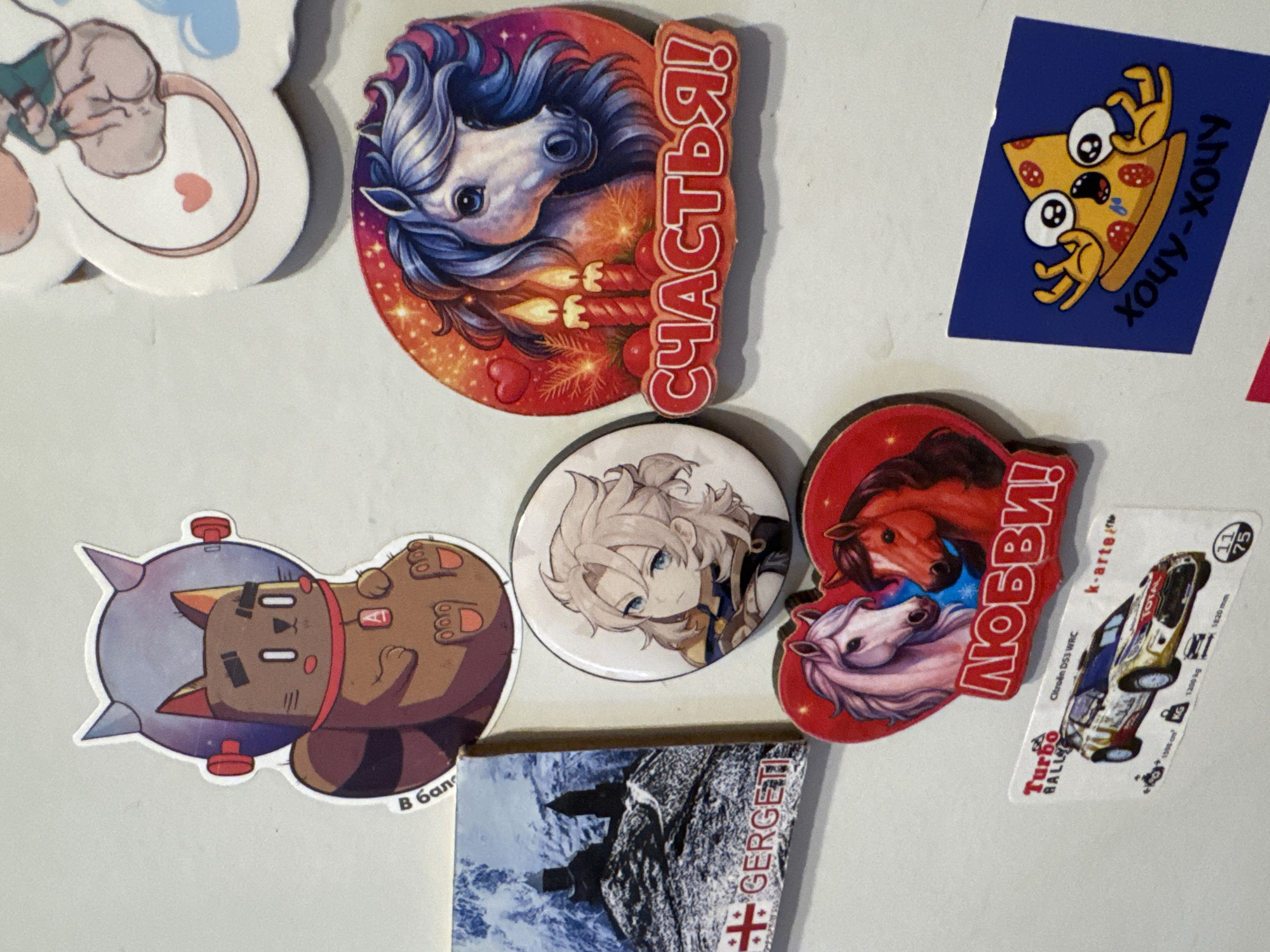 The image shows a white, textured surface, likely a wall, adorned with a collection of magnets and stickers.
At the top left is a sticker of a brown cartoon cat wearing a silver astronaut helmet with red accents. The cat has a red collar with a red 'A' pendant. Text in white Cyrillic is partially visible on the left side of the sticker, reading "В Бан".
To the right of the cat sticker, there is a large circular magnet featuring a stylized drawing of a white horse with a blue mane, set against a vibrant orange and pink background with sparkles. Two red candles and red berries are depicted in front of the horse. The Russian word "СЧАСТЬЯ!" (Happiness!) is written in red, block letters below the horse.
Below the astronaut cat sticker is a square magnet depicting a snowy mountain landscape with two dark buildings on a peak. The word "GERGETI" is written in red text along the bottom edge of this magnet, with a red Georgian cross symbol to its left.
Center right is a round magnet featuring a character with light blonde hair and blue eyes, identified as Albedo from the game Genshin Impact. The character has a serious expression and is wearing a golden and black outfit.
Below the Albedo magnet and to the left of the horse magnet is a heart-shaped sticker with two cartoon horses, one pink and white and the other reddish-brown, looking at each other. The Russian word "ЛЮБВИ!" (Love!) is written in red, block letters below the horses.
Further down and to the left is a rectangular sticker of a yellow and white rally car, identified as a "Citroen DS3 WRC" and a "Turbo Rally" sticker. It displays technical specifications like "1598 cm³", "1200 kg", and "1820 mm". The "k-arte" logo is also visible.
At the bottom right is a square blue sticker with a cartoon slice of pizza with wide, startled eyes and arms. Below the pizza, the Russian phrase "хочу-хочу" (I want-want) is written in black, cursive letters.
The image is a close-up view, and the exact location within Яромінскі сельскі Савет, Belarus is not specified by any visual cues in the image itself. The surface appears to be clean and well-lit.