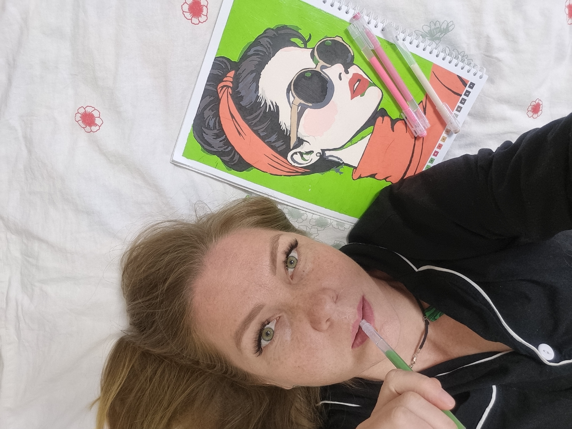 A fair-skinned woman with freckles and green eyes is lying on a white bed sheet with small red flower patterns. She is wearing a black pajama top with white trim. She holds a green pen between her lips and her right hand holds the end of the pen. To her left is a notebook with a spiral binding, on which is a pop art style portrait of a woman wearing sunglasses and a red scarf. Two other pens, one pink and one beige, are lying on top of the notebook. The scene is photographed from an overhead angle. While the location is not evident from visual details of the image, the prompt specifies the city of Chaikovsky, Russia.