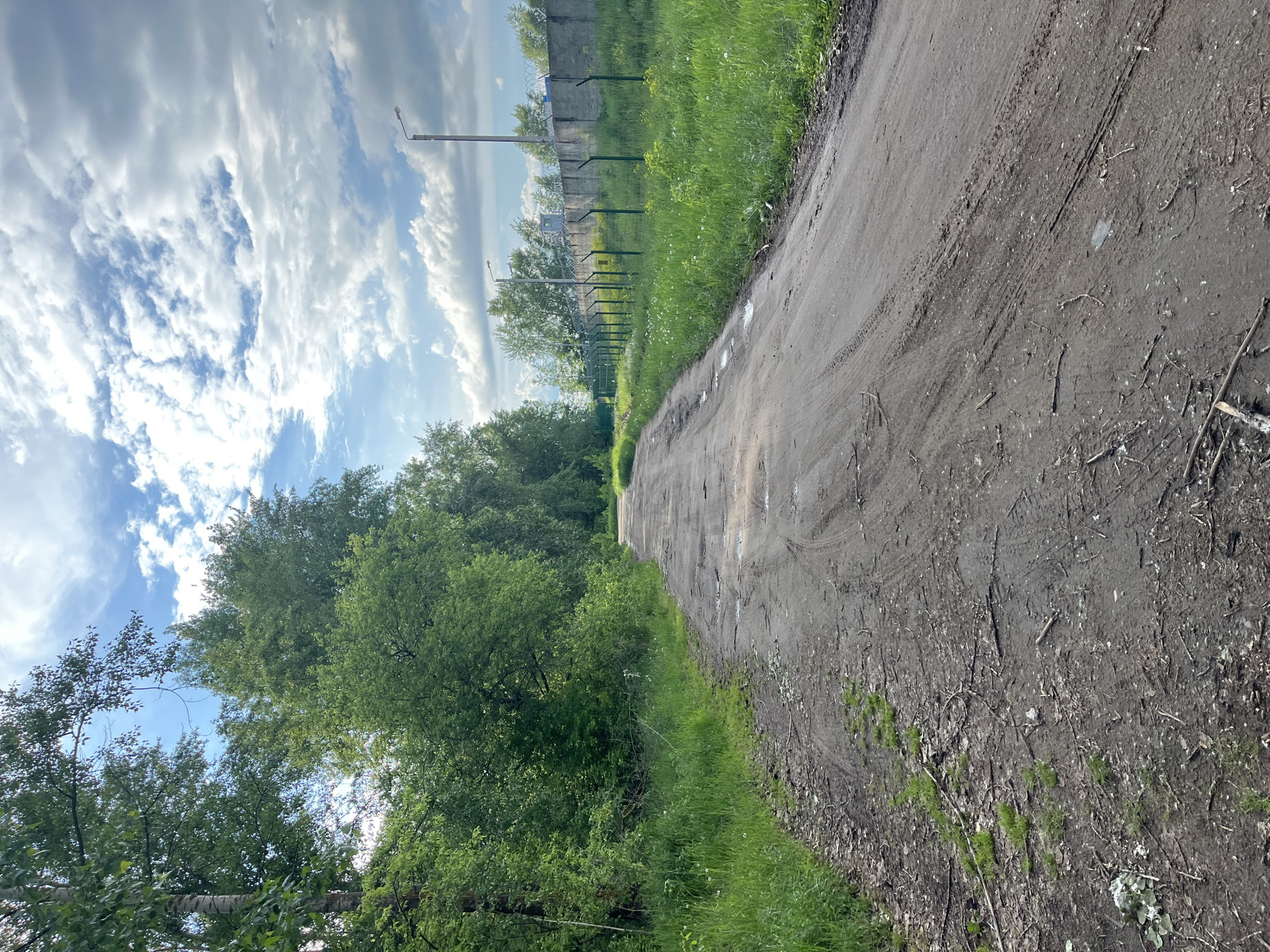 The image depicts a dirt road leading into a wooded area in Chaikovsky, Russia. The road surface is uneven and shows signs of vehicle traffic. To the left of the road is a dense line of trees and vegetation. To the right, a grassy area borders the road, with a concrete fence and lampposts visible further in the distance. The sky is partly cloudy with patches of blue sky.