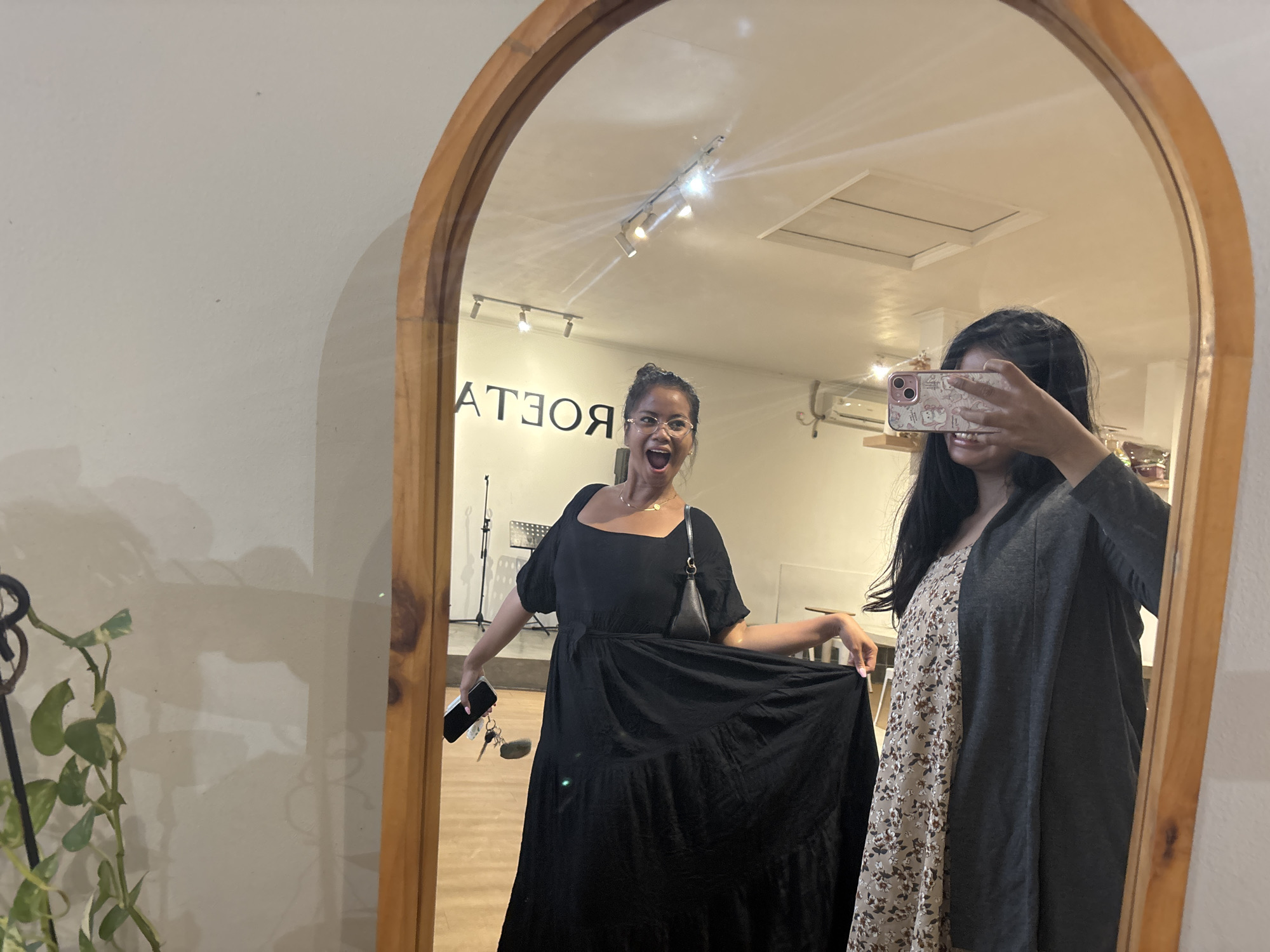 The image shows two women reflected in an oval-shaped mirror with a wooden frame. One woman stands within the reflection, wearing a black square-neck maxi dress and holding the hem out to the side; she has an open mouth expression and glasses, with a purse over her shoulder and keys in hand. The second woman is partially visible in the reflection, wearing a floral dress and a grey cardigan, holding a cell phone up to take the picture. The environment appears to be an indoor space with a white ceiling and recessed lighting. A plant is partially visible in the foreground to the left of the mirror. Backwards writing on the wall can be seen in the background in the reflection. The photo was taken in North Jakarta, Indonesia.