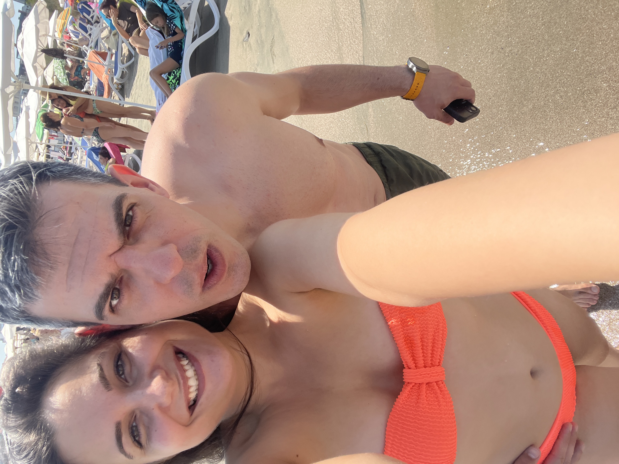 A man and a woman are taking a selfie on a beach in Pomorie, Bulgaria. The man is shirtless and wearing green shorts, and the woman is wearing an orange bikini. They are both smiling and looking at the camera. In the background, other beachgoers are visible relaxing on lounge chairs under umbrellas. The sand is light brown and the water is not visible. The overall atmosphere is one of leisure and enjoyment on a sunny day.