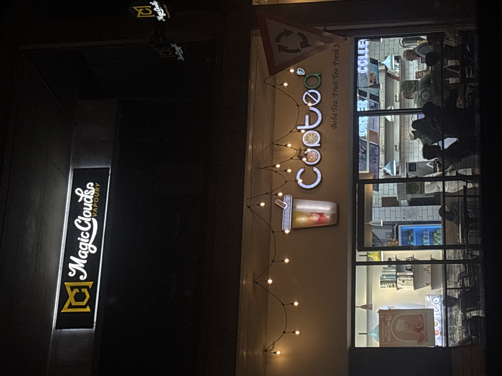 The image presents a night scene in what appears to be a commercial district, potentially in Durban, South Africa, given the signs in the image. On the left, a vertical neon sign reads "Magic Clouds Vapoury" with an icon above the text. To the right, the storefront of a "CooToo" boba tea shop is visible, decorated with string lights and featuring a logo with a stylized drink. The boba tea shop sign reads "Boba Tea Fruit Tea Fresh". Through the shop's windows, silhouettes of people are visible inside. Inside the shop, there are shelves with what appear to be fruits and drinks, along with advertisements and books. The lighting is dim, emphasizing the illuminated signs and store interior. A traffic sign with an arrow is seen on top of the boba tea store sign.