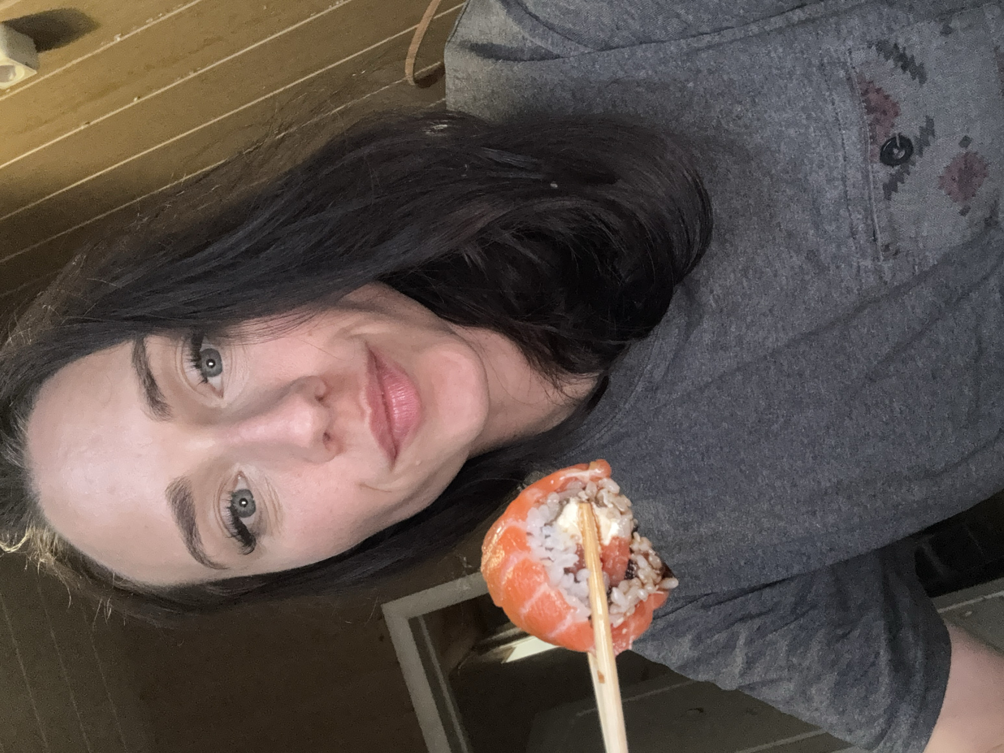 A woman is holding a piece of sushi with chopsticks in front of her face, taking a selfie in what appears to be a room with wooden paneled walls. The woman has dark hair, fair skin, and is wearing a gray t-shirt. The sushi is a roll with salmon on the outside and rice and filling inside. The background is out of focus but shows a doorway and the wooden walls. The lighting is dim. The image appears to have been taken indoors. There is no indication of location in Saint Petersburg, Russia.