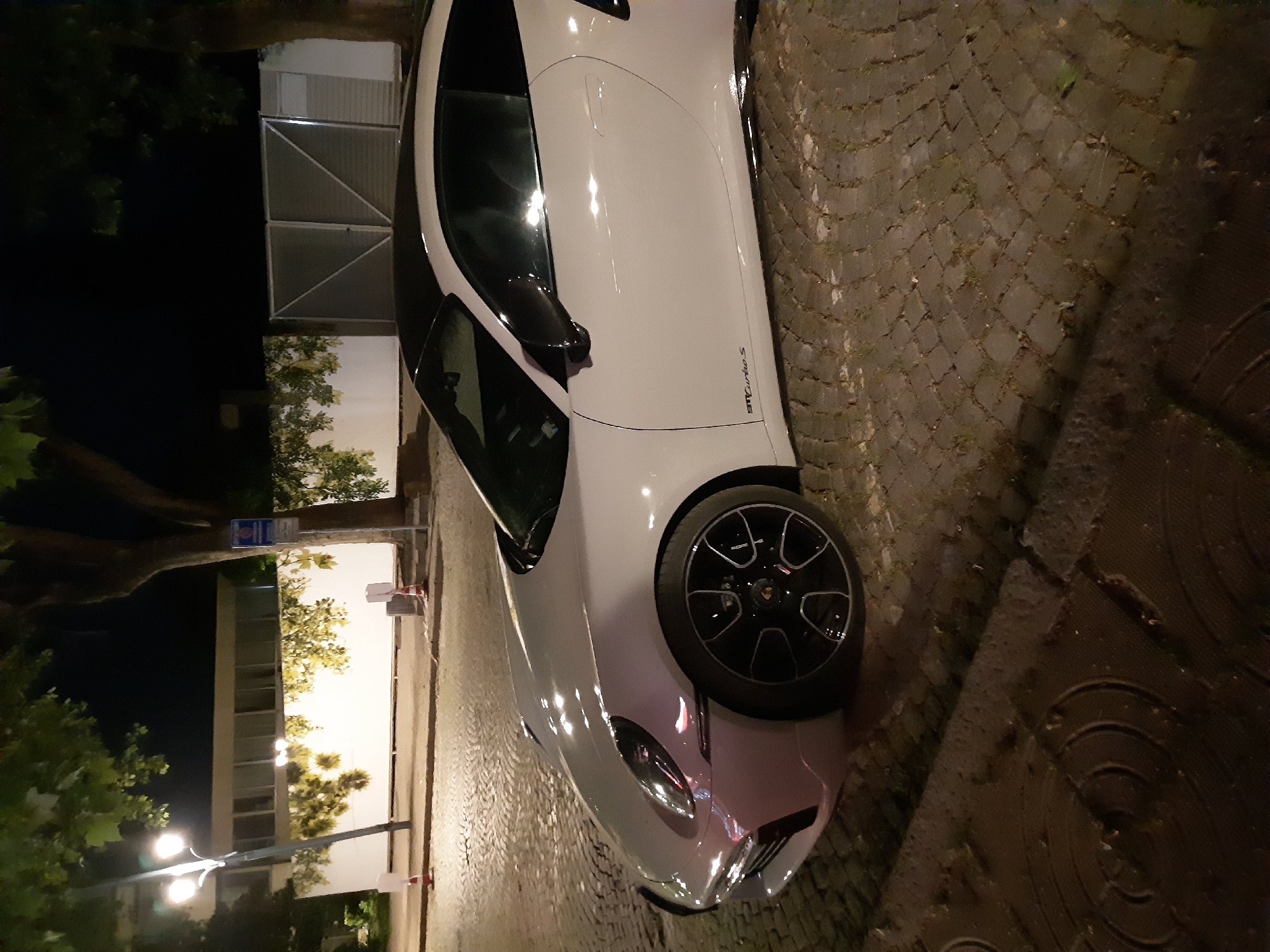 A white Alfa Romeo 4C Spider is parked on a cobblestone street in Plovdiv, Bulgaria. The car is positioned at an angle, with the front end closer to the viewer. To the left of the car, there is a building illuminated by streetlights and trees. The background is dark, and the street is wet, possibly from rain. The car's "GT Turbo S" branding is visible.