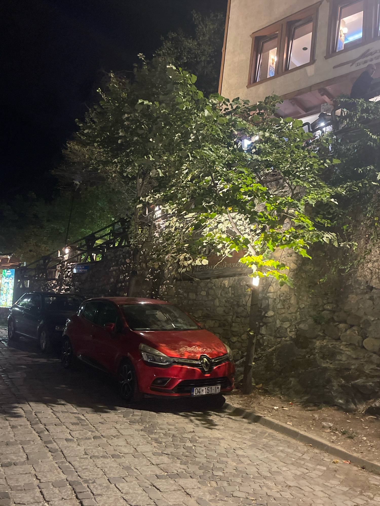 The image depicts a nighttime scene on a cobblestone street in Prizren, Kosovo, in front of a building with a stone wall. A red Renault Clio is parked on the street, facing left, with its license plate visible as "04-151-IM". Behind it, a dark-colored car is also parked. Lush green trees with illuminated leaves are prominent in the foreground and frame the building, casting shadows on the street. The building itself has multiple windows, some of which are lit from within, suggesting it is a restaurant or establishment. A lamppost illuminates the area near the trees. The street appears to be a pedestrianized or low-traffic area, typical of historical city centers. The overall atmosphere is calm and serene, illuminated by artificial lights against the dark night sky.