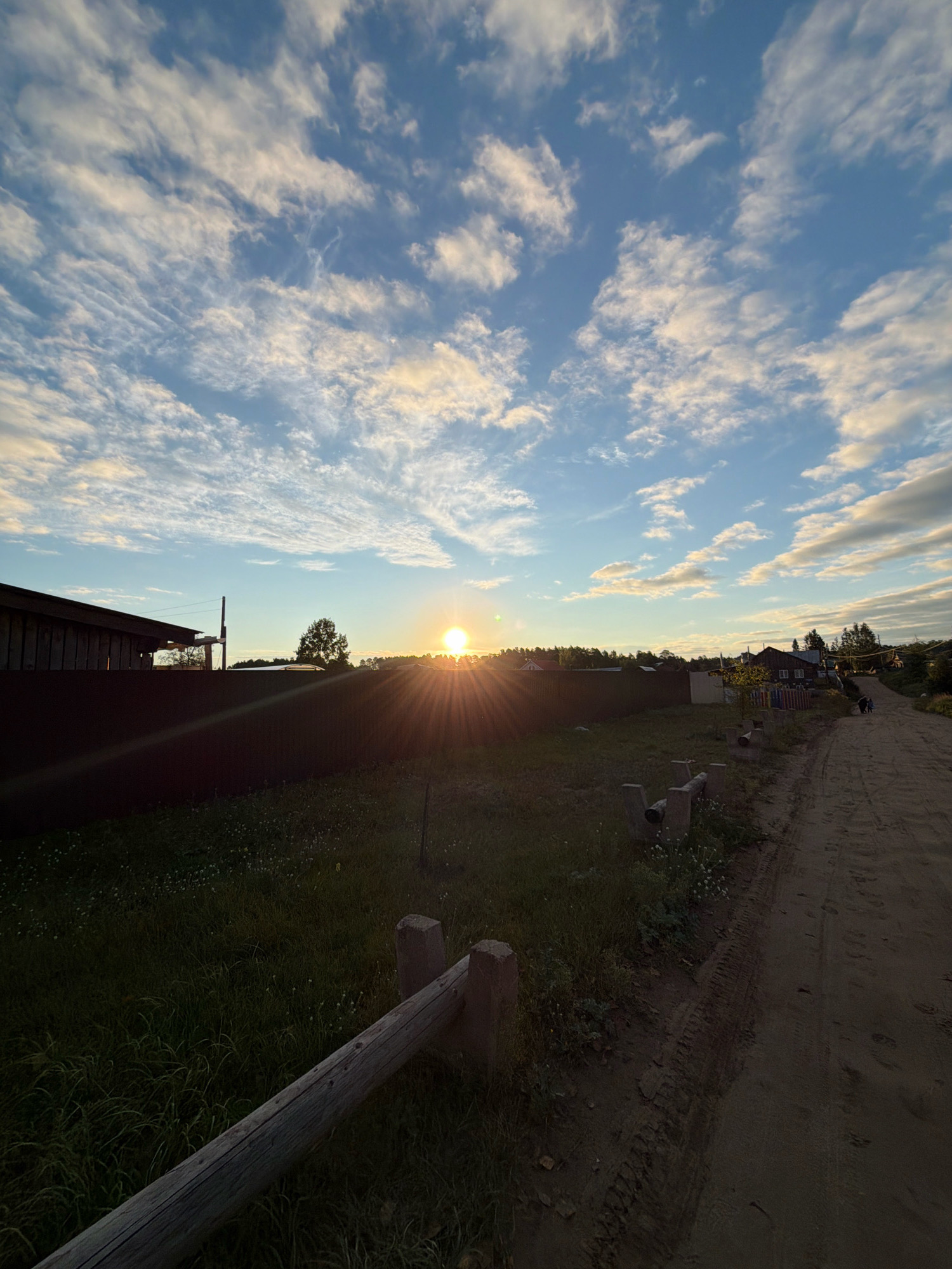 The image depicts a rural landscape at sunset, likely in the outskirts of Chaikovsky, Russia. The sun is low on the horizon, casting a warm glow and creating a lens flare effect. The sky is partly cloudy with scattered white and golden clouds against a blue backdrop. In the foreground, a dirt road runs diagonally from the right to the left. Adjacent to the road is a grassy field with small white flowers and a weathered wooden fence. To the left of the field, a dark wooden fence runs horizontally, with the side of a building visible behind it. In the middle ground, several houses with visible windows and roofs are clustered together. Trees are scattered throughout the scene. Two figures, appearing to be people, are walking on the dirt road in the distance. The overall atmosphere is serene and tranquil, characteristic of a rural setting at dusk.