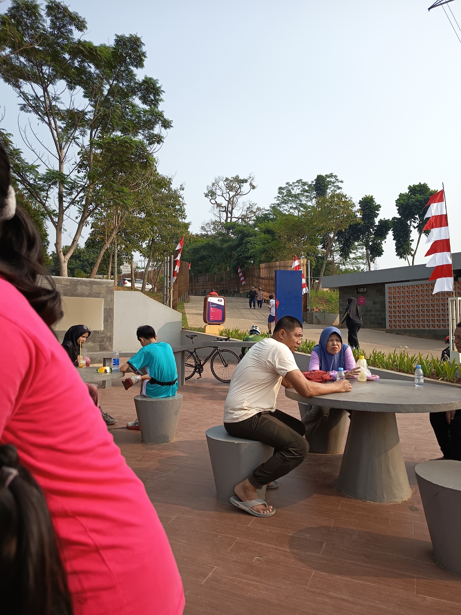 The image depicts an outdoor seating area at Taman Lapangan Banteng in East Jakarta, Indonesia, during daylight. Several people are present, engaged in various activities. In the foreground, a person wearing a bright pink top is partially visible from behind, with their back to the camera. To the left, a woman in a black hijab and a woman in a blue hijab are seated at a circular concrete table, with drinks in front of them. To the right of the table, a man in a light-colored t-shirt and dark pants is seated, facing the women, appearing to engage in conversation. Another man is seated to the far right, his face obscured. In the middle ground, a person in a teal shirt sits on a concrete stool. A bicycle is parked nearby. The environment features trees and foliage, with red and white Indonesian flags visible in the background, as well as architectural elements of the park, including concrete structures and a building with patterned walls. The ground is paved with reddish-brown tiles. The sky is clear and bright.