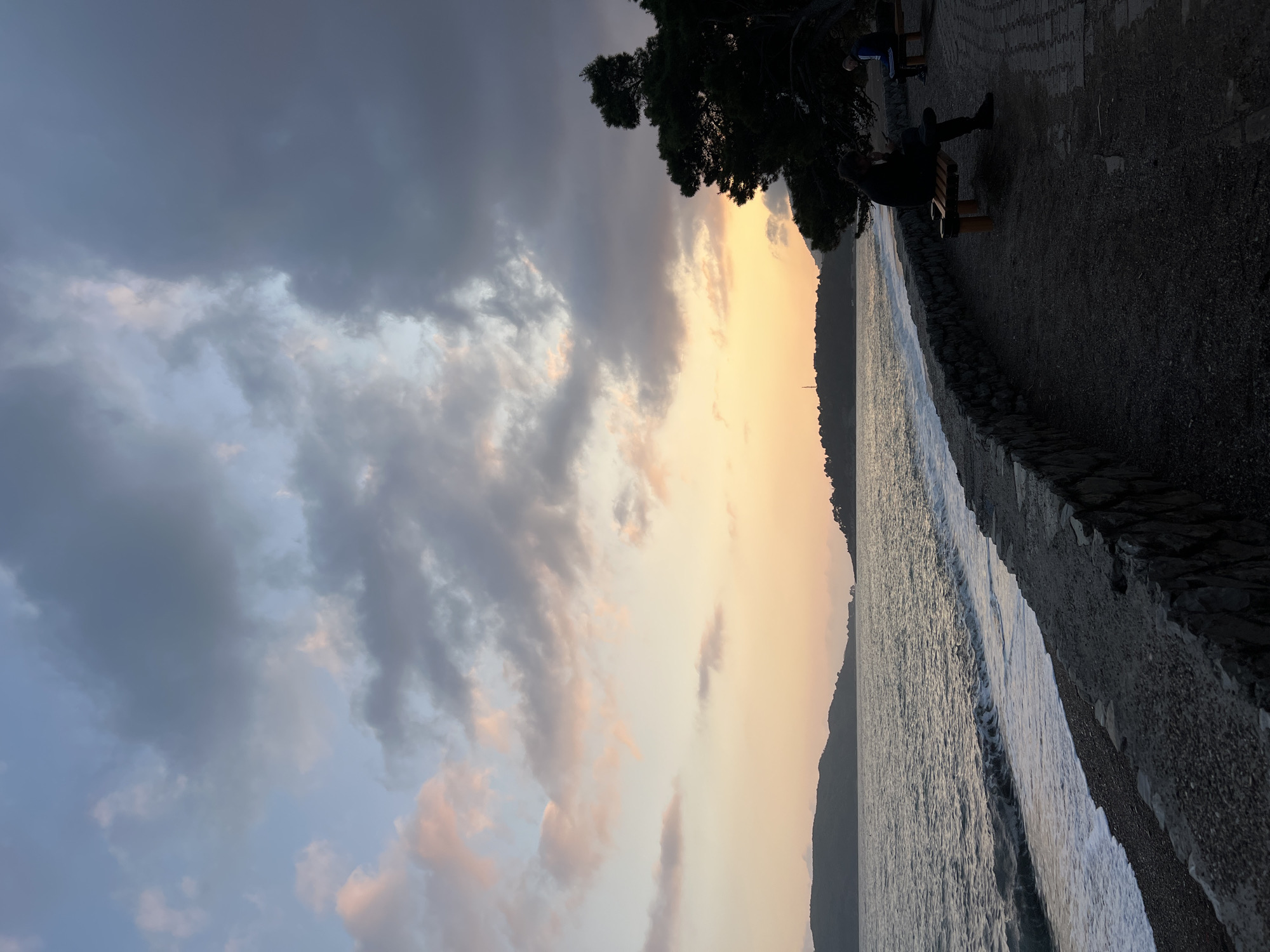This image captures a tranquil coastal scene in Rafailovići, Montenegro, at sunset or dusk. The sky, dominating the upper portion of the frame, displays a dynamic blend of colors and cloud formations. Dark, heavy gray and blue clouds are prominent on the left, suggesting a recent or impending weather shift, while towards the right horizon, the sky glows with warm oranges and soft yellows, indicative of the setting sun. Scattered, lighter clouds catch this warm light.
Below the sky, the sea extends horizontally, with gentle waves creating white foam as they break along the shore. The water surface appears a muted, reflective gray. A low, stone-stacked wall borders the sea, separating it from a dark, gravelly or paved path.
In the foreground on the right, a person is seated on a wooden bench, their figure largely silhouetted against the lighter horizon. Another bench is partially visible further back. A dense, dark green tree with intricate branches frames the upper right corner, contributing to the shadowy foreground. A distant, dark mountainous landmass or coastline stretches across the horizon, silhouetted against the warm sky. The overall mood is peaceful and contemplative, enhanced by the dimming light. No visible text is present in the image.