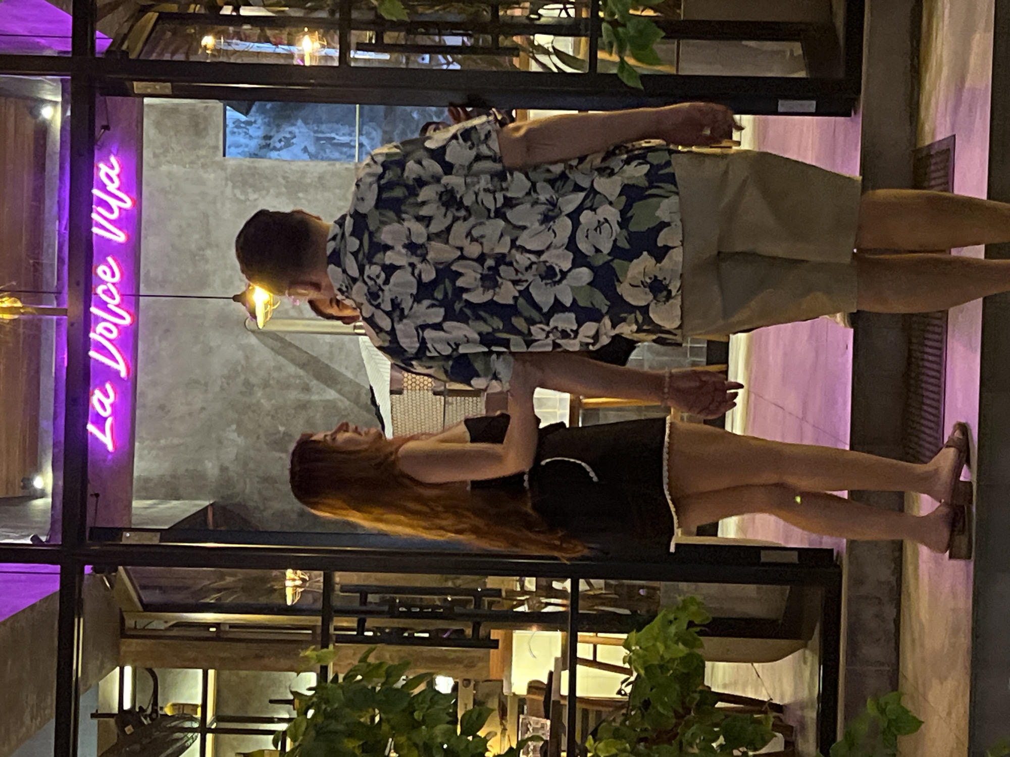A couple stands at the entrance of a building in Ubud, Indonesia. The woman, on the left, has long, auburn hair and wears a black strapless dress with white trim. She faces left, talking to the man. The man, on the right, wears a short-sleeved, floral-patterned shirt and khaki shorts. He faces away from the camera, looking into the building. Behind them, a neon sign glows in pink, reading "La Dolce Vita". The entrance is framed by a dark metal structure with glass panes. Green plants are visible on the left side of the frame. The ground is paved with light-colored tiles, and a dark mat lies at the entrance. The lighting suggests it is evening.