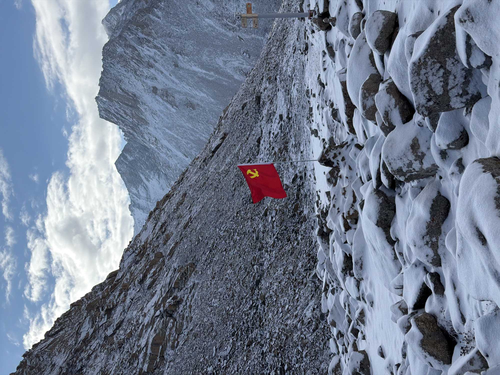 The image displays a rugged, snow-covered mountainous landscape under a partially cloudy blue sky. The scene is dominated by steep, rocky slopes dusted with snow, suggesting a high-altitude and cold environment.
In the mid-ground, a vibrant red flag featuring a yellow hammer and sickle emblem is planted, standing out against the muted tones of the mountain. The flag is attached to a pole that is partially obscured. To the right of the flag, a metal pole with several small, indistinct signs or markers attached near its top is visible.
The foreground on the right consists of numerous large, dark rocks, heavily blanketed with fresh, white snow, creating interesting textural patterns. Beyond the flag, the terrain continues to be rocky and snow-dusted, with the contours of distant peaks visible against the sky. The sky is bright blue with scattered white, elongated clouds.
Given the snow coverage and the nature of the terrain, the weather appears to be cold, and the time of day is likely late morning or early afternoon, indicated by the bright, diffused light. There are no people visible, and no specific activity is taking place other than the flag standing proudly in the harsh environment. The only visible "text" is the hammer and sickle symbol on the flag; no readable words are present on the flag or the signs on the pole. The presence of the flag, particularly a communist one, in a mountainous region of what is given as Parga town, China, suggests a significant or marked location within the area.