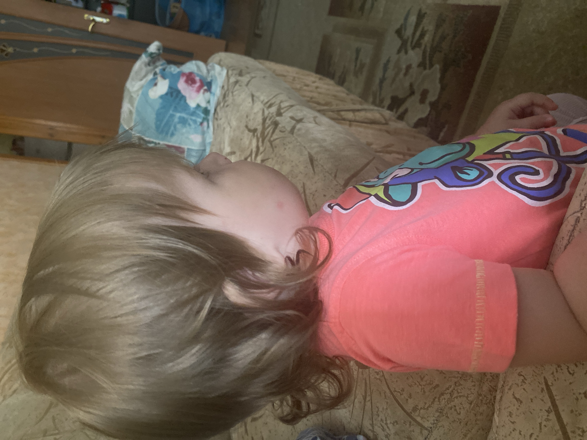A young child, seen from the side, is sitting on a couch. The child has light brown, wavy hair. The child is wearing a bright pink t-shirt with a colorful, abstract design on the front. The background shows a couch with a textured beige fabric, a pillow with a floral pattern, and a doorway with a wooden door and a gold handle. A patterned rug is visible on the floor. The scene appears to be indoors, possibly in a living room or common area.