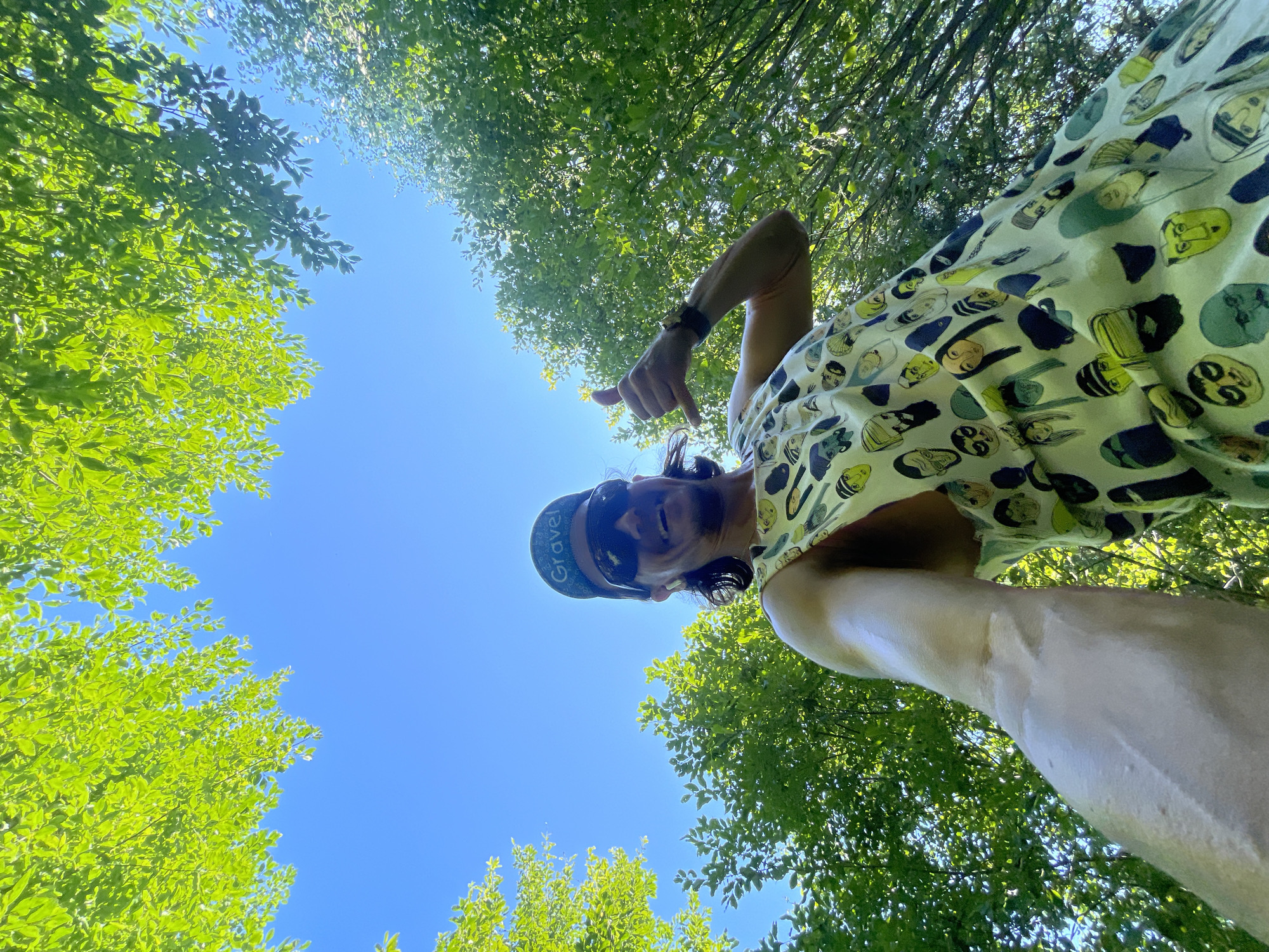 An outdoor, low-angle, eye-level selfie shows a smiling man wearing a patterned sleeveless shirt with faces and a baseball cap with "Gravel" written on it, sunglasses, and an earbud. He is giving a thumbs-up and a shaka hand gesture. The man is outdoors surrounded by lush green trees and a bright blue sky. The location is the city of Perevoznoye, Russia.
