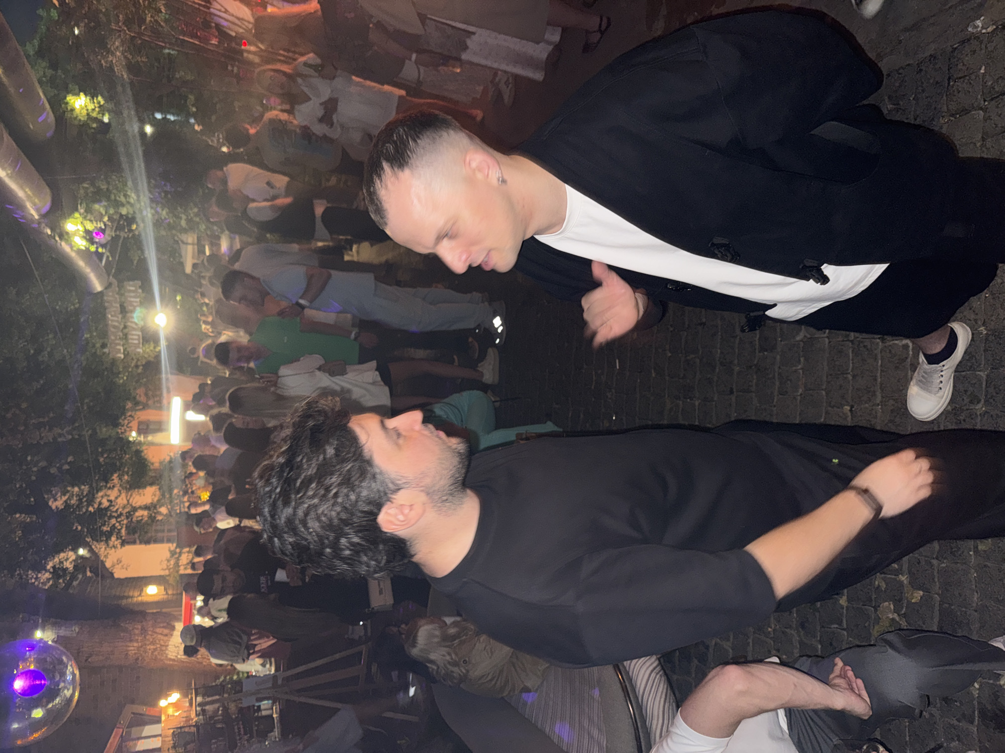 A medium-shot, full-color, outdoor, nighttime photograph shows two men facing each other and engaged in conversation. Both men are the primary subjects and are positioned in the foreground, slightly off-center. The man on the left has dark, wavy hair, a beard, and is wearing a black t-shirt. He is looking towards the man on the right. The man on the right has short, dark hair with a buzz cut, a white t-shirt, and a black open-front jacket. He is gesturing with his right hand and looking at the man on the left.
The environment is a crowded outdoor venue, likely a bar or club with an urban feel. The ground is paved with cobblestones, suggesting a historic or pedestrianized area. In the background, numerous people are visible, some standing, some sitting, and some moving around, creating a lively atmosphere. The lighting is dim, with some artificial lights illuminating the scene, including string lights overhead and lamps, casting a warm glow. A disco ball is visible in the upper left corner, reflecting light. The overall impression is of a social gathering or event at a city nightlife establishment. The image is not specifically identifiable as being in Moscow, Russia, or any other specific city without further context or identifying landmarks.