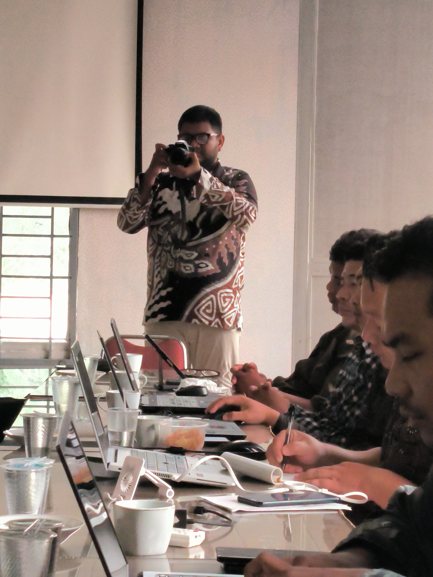 The image captures a group of individuals in a room, likely a classroom or meeting space, in Lhokseumawe, Indonesia. A man in the foreground, wearing glasses and a patterned shirt, stands and holds a camera to his eye, seemingly taking a photograph of the attendees. In front of him, several people are seated at a table, engrossed in their work, with laptops, notebooks, and cups spread out before them. The room is brightly lit, with natural light streaming in from a window on the left side of the frame. The overall atmosphere suggests a productive gathering, possibly a workshop or lecture, in the city.