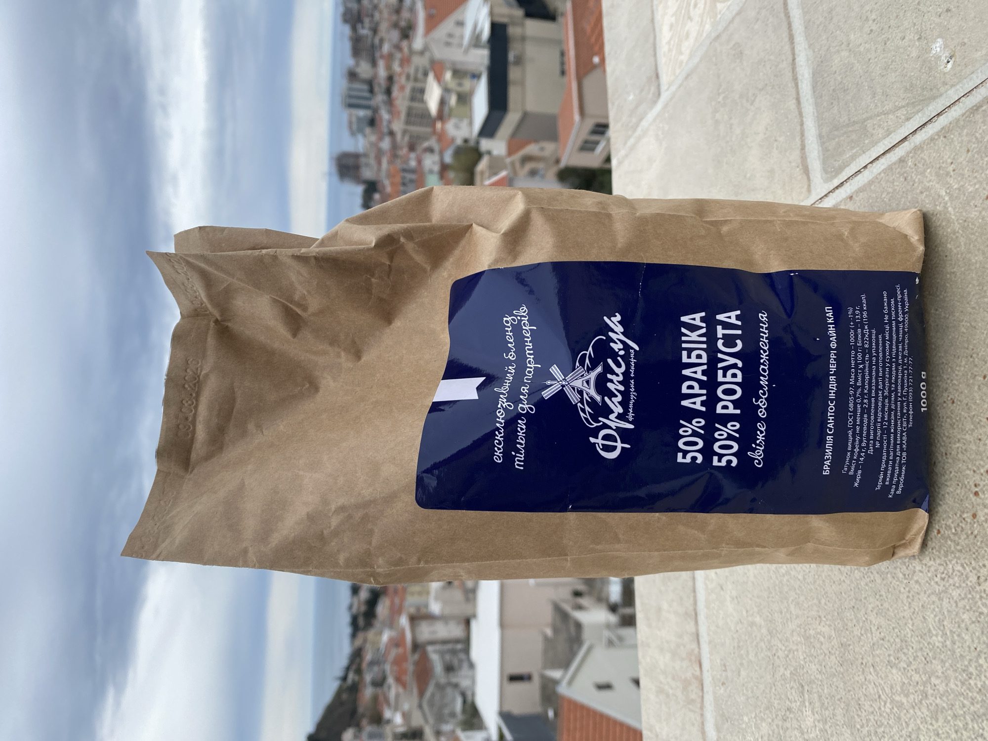 A bag of coffee sits on a balcony overlooking the cityscape of Budva, Montenegro. The bag is brown paper with a blue label that reads "50% Arabica, 50% Robusta." The background shows buildings with red-tiled roofs typical of the area, stretching towards the sea under a cloudy sky. The balcony has a light tan surface with mottled patterns.