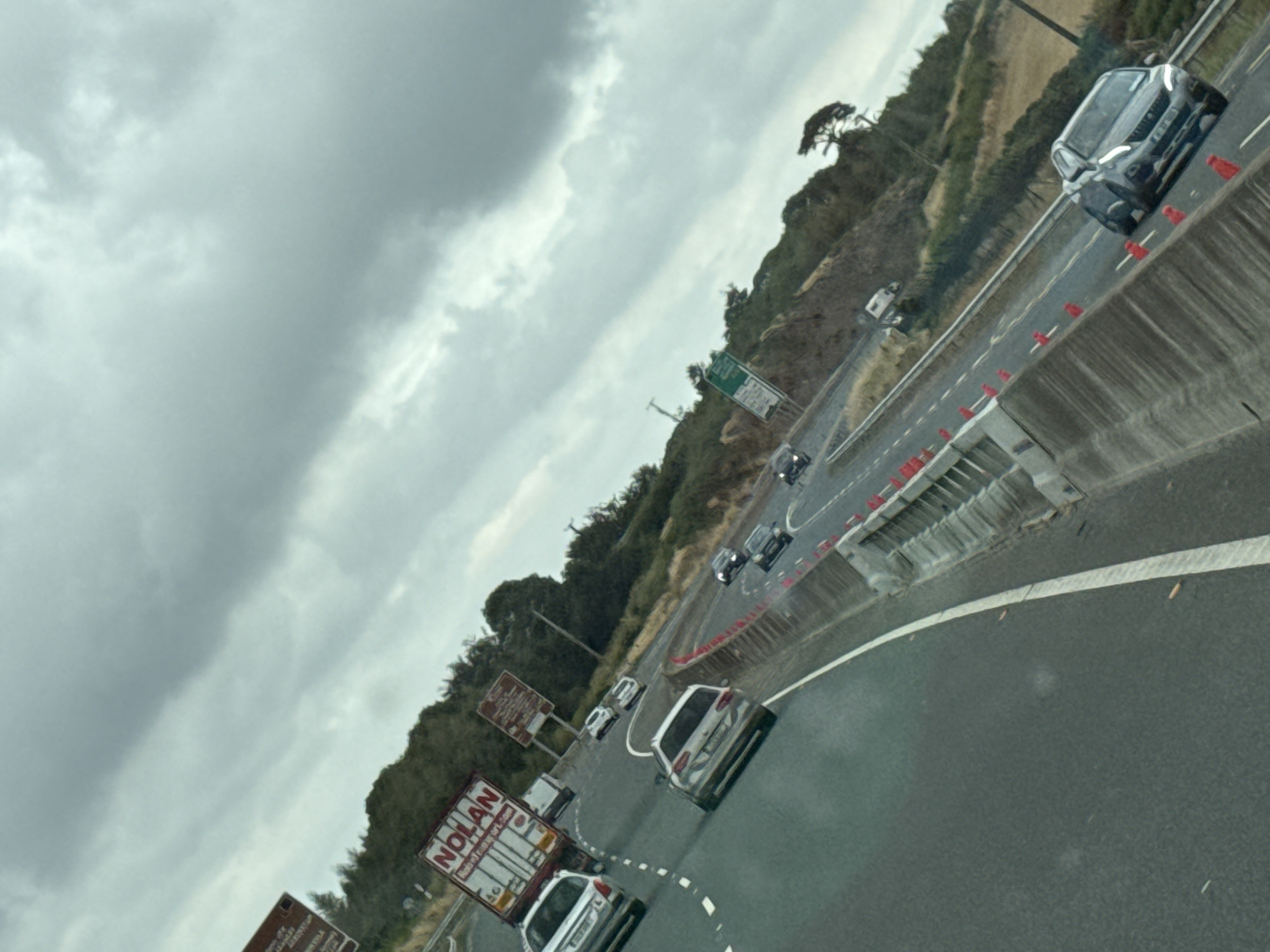 A grey, overcast sky looms over a multi-lane highway in the New Ross Municipal District, Ireland. The highway curves to the right, with a concrete barrier on the right side of the road, adorned with red traffic cones. Several vehicles are visible on the highway, including a white truck with "NOLAN" written on its side, a white car, a silver station wagon, and a dark-colored SUV. The surrounding landscape features a wooded hillside on the right and a grassy embankment on the left. A street light is visible on the left side of the road. The image is taken from a moving vehicle, as indicated by the motion blur and the angle of the shot.